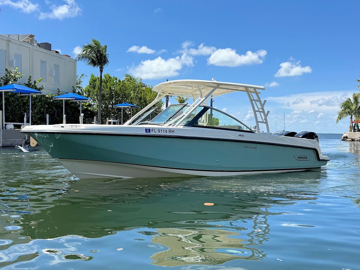 Boat in teal and white, docked on calm water with buildings and blue sky in the background.