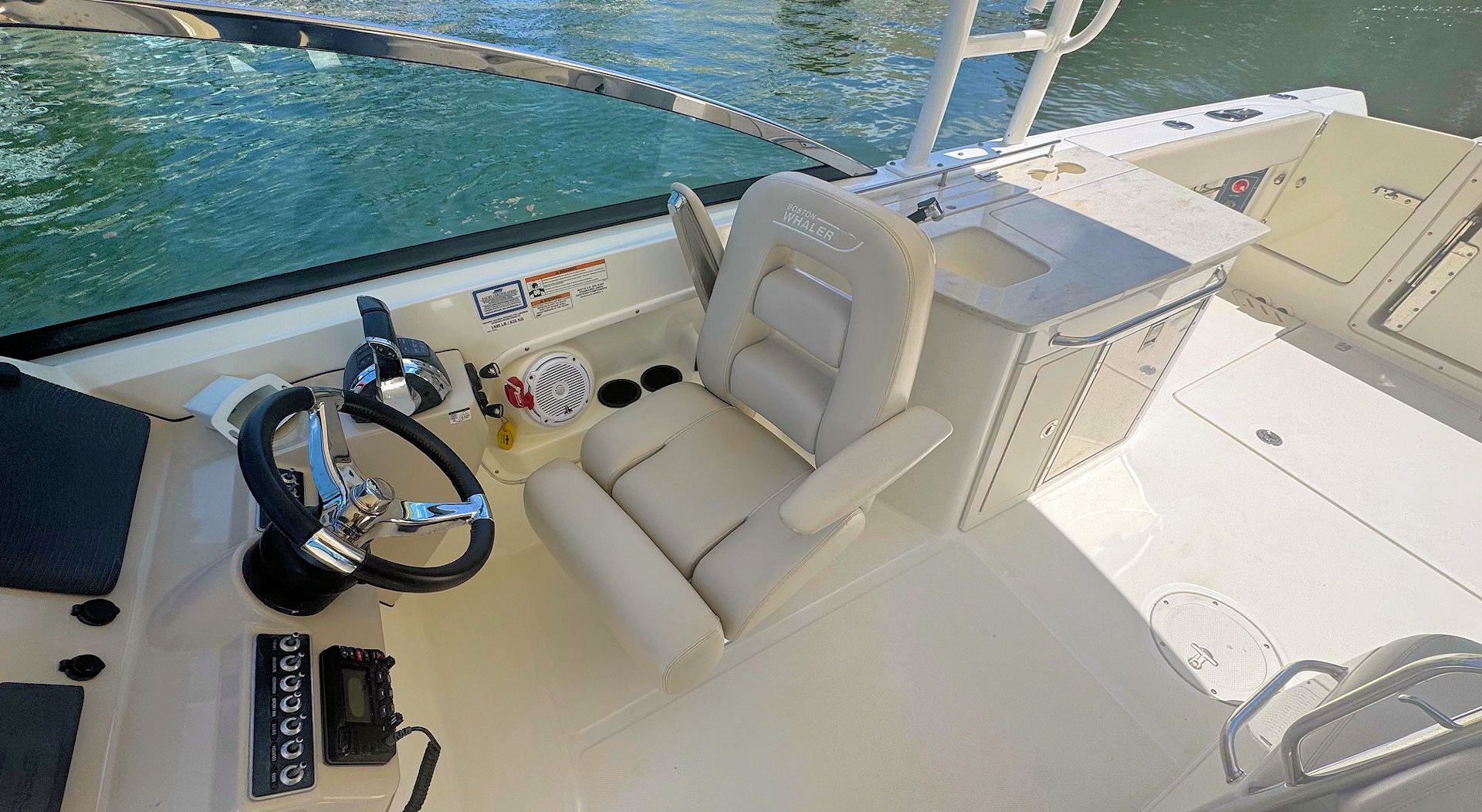 The helm of a boat featuring a steering wheel, beige captain's chair, and a console with a sink on a white deck.