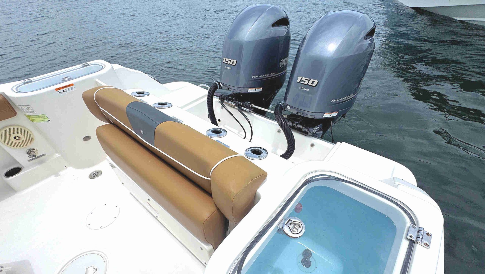 Boat stern with two gray outboard motors and tan seat cushions beside a small water hatch on calm water