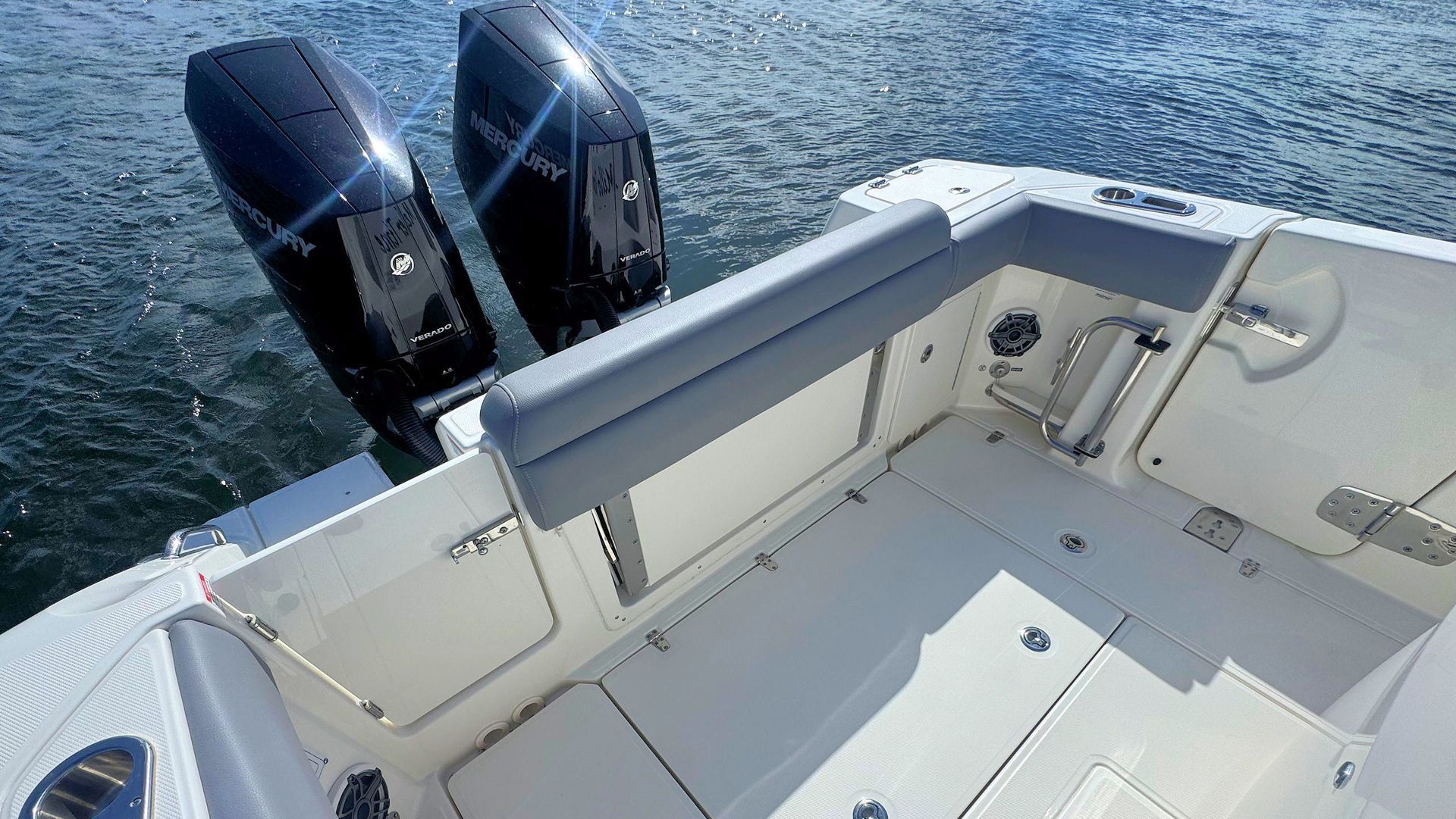 The stern of a boat with two Mercury outboard motors, a grey cushioned bench seat, and a white deck on the water.