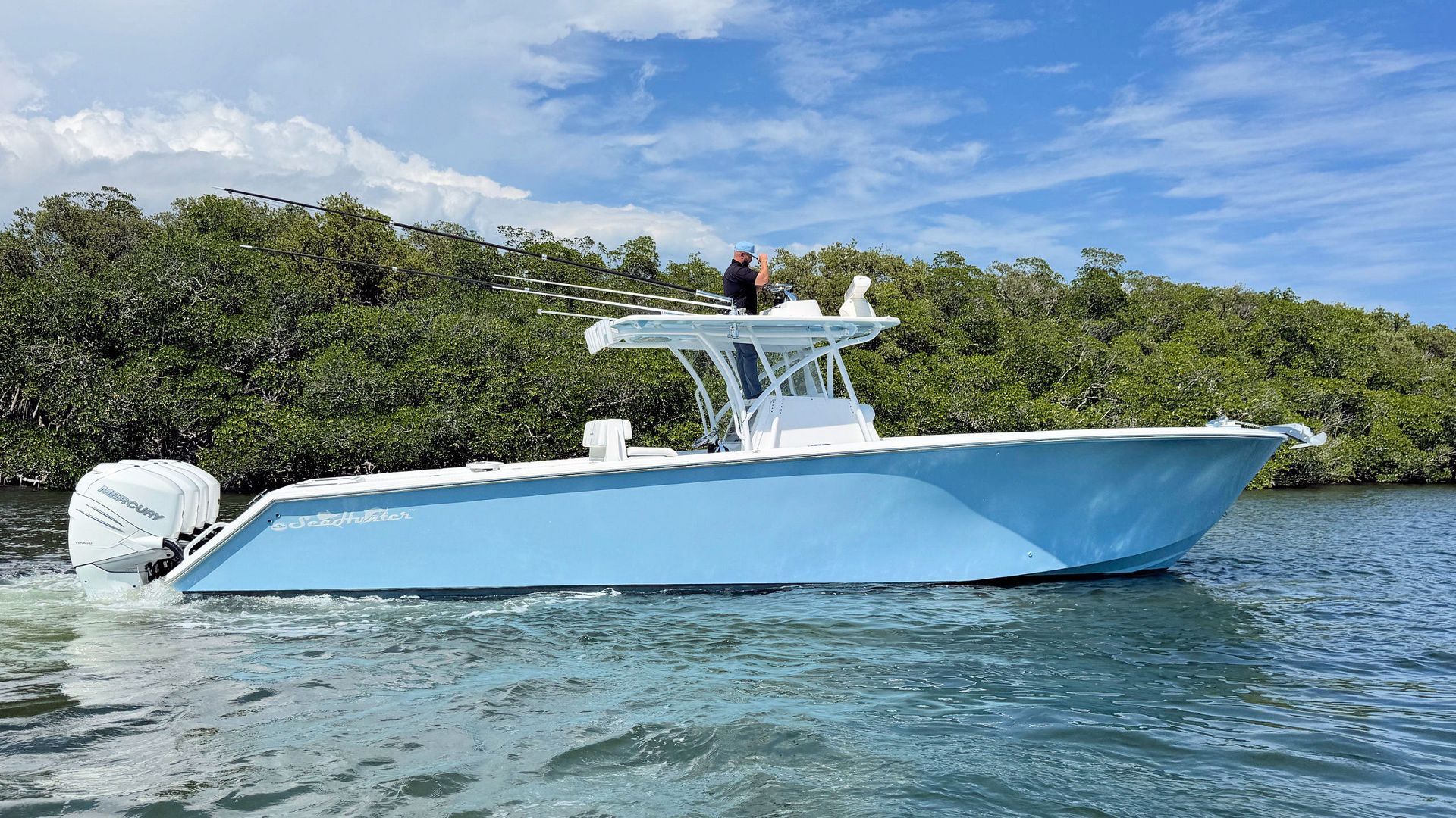 Light blue center console boat cruising on calm water near a green, tree-lined shore under a blue sky