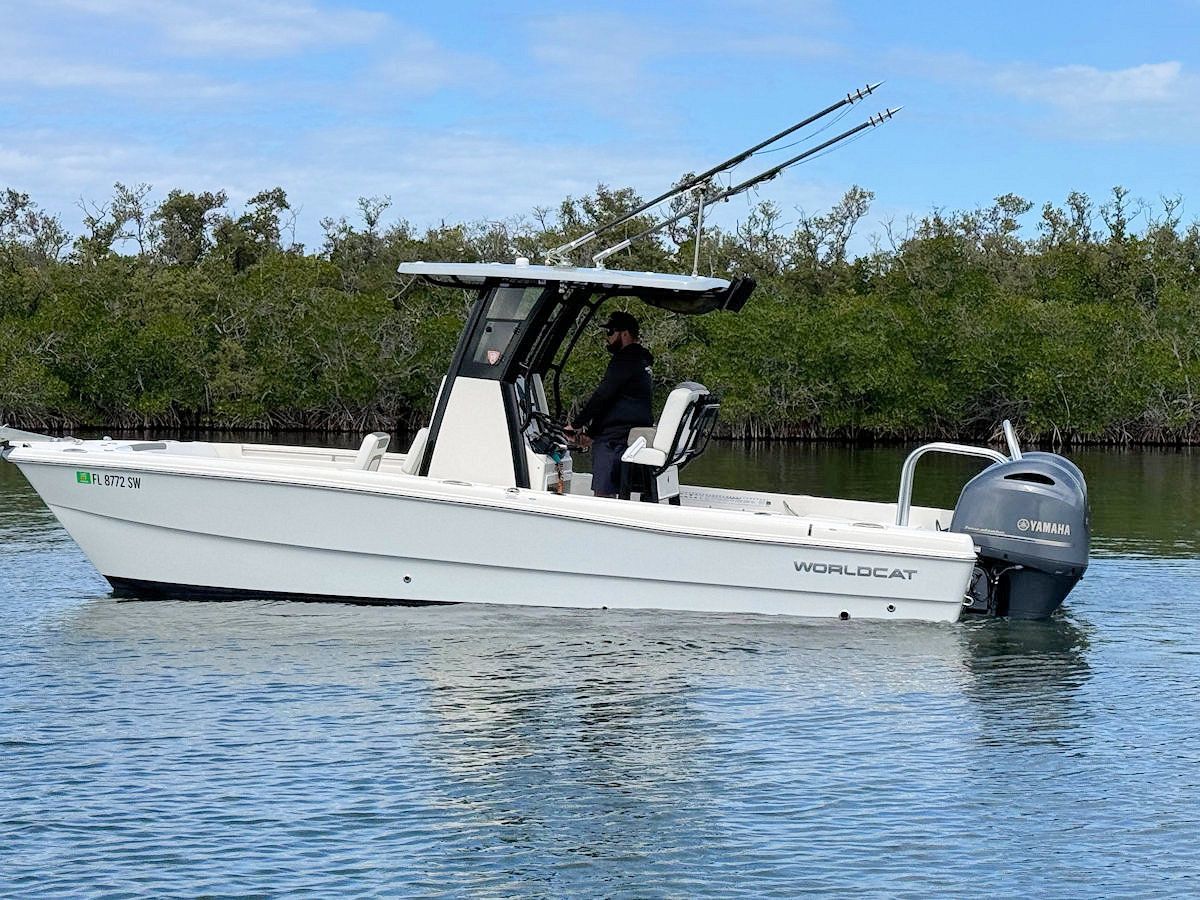 White motorboat on water with a person at the helm, trees in the background.