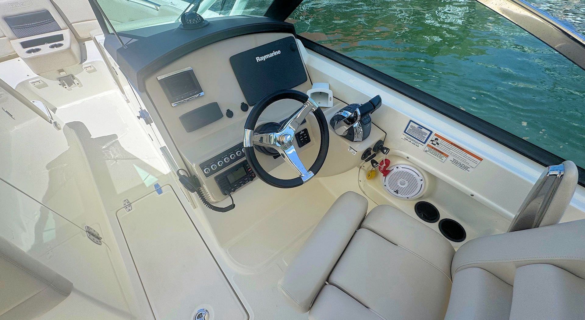 The cream-colored helm of a boat features a steering wheel, navigation display, and beige cushioned seating.