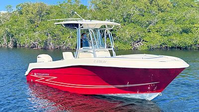 Red and white motorboat on water, with a person at the helm, in front of lush green trees.