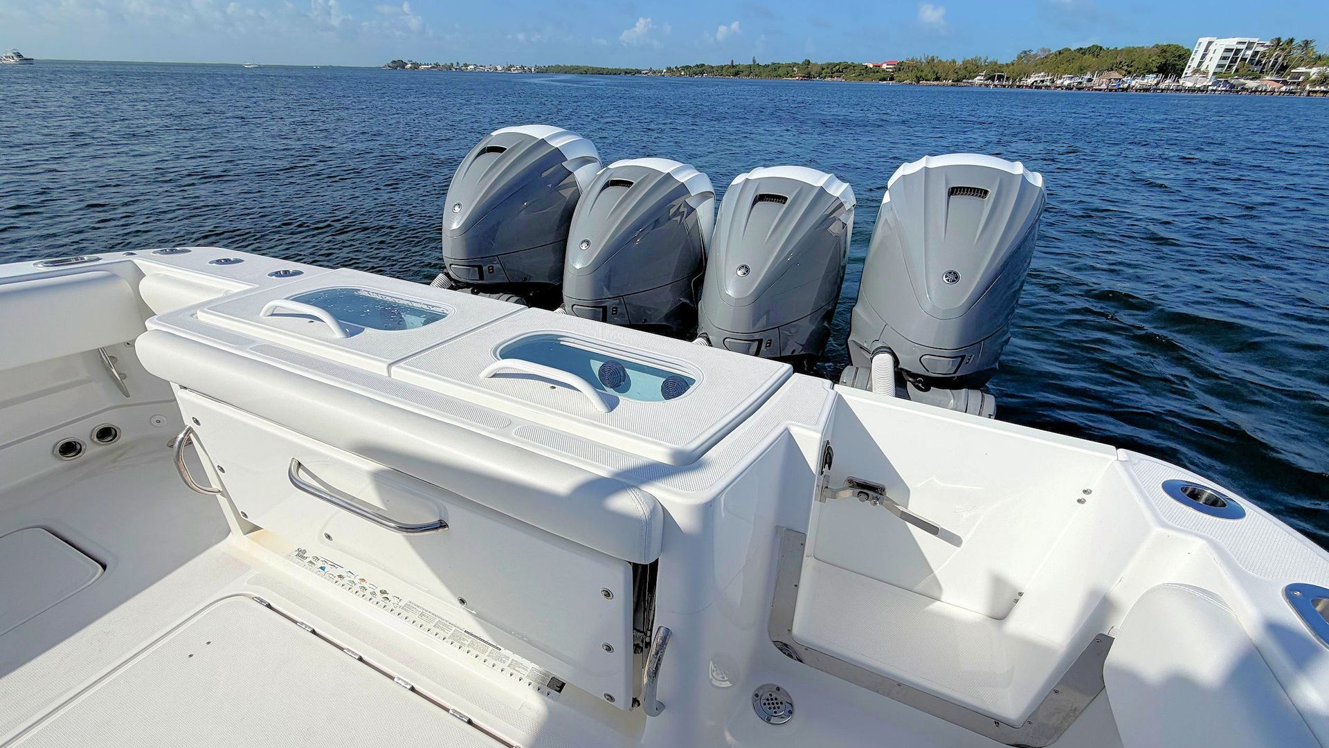 White boat deck with four outboard motors on open blue water near a shoreline.