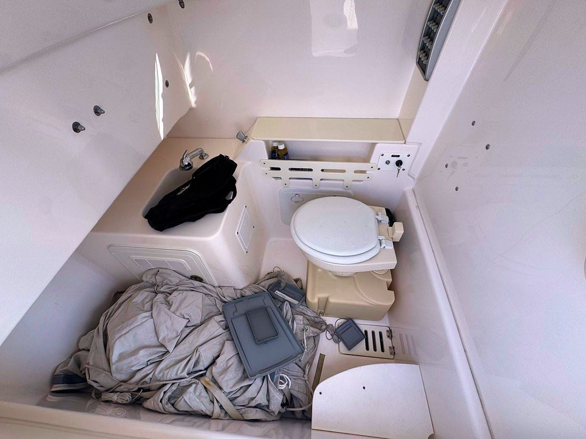 Interior of a small boat, likely a head. White walls surround a toilet, a sink, and storage area containing items.
