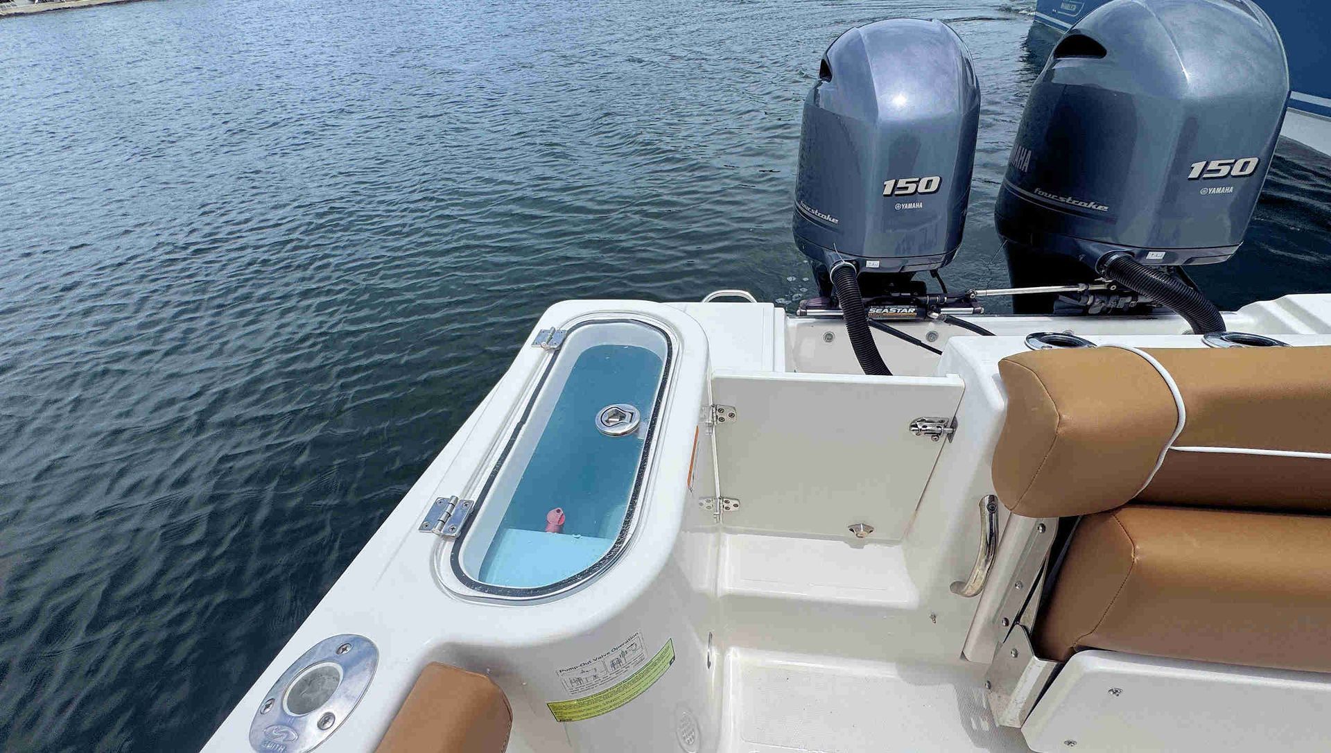 Boat stern with two gray outboard motors and tan seat cushions beside a small water hatch on calm water