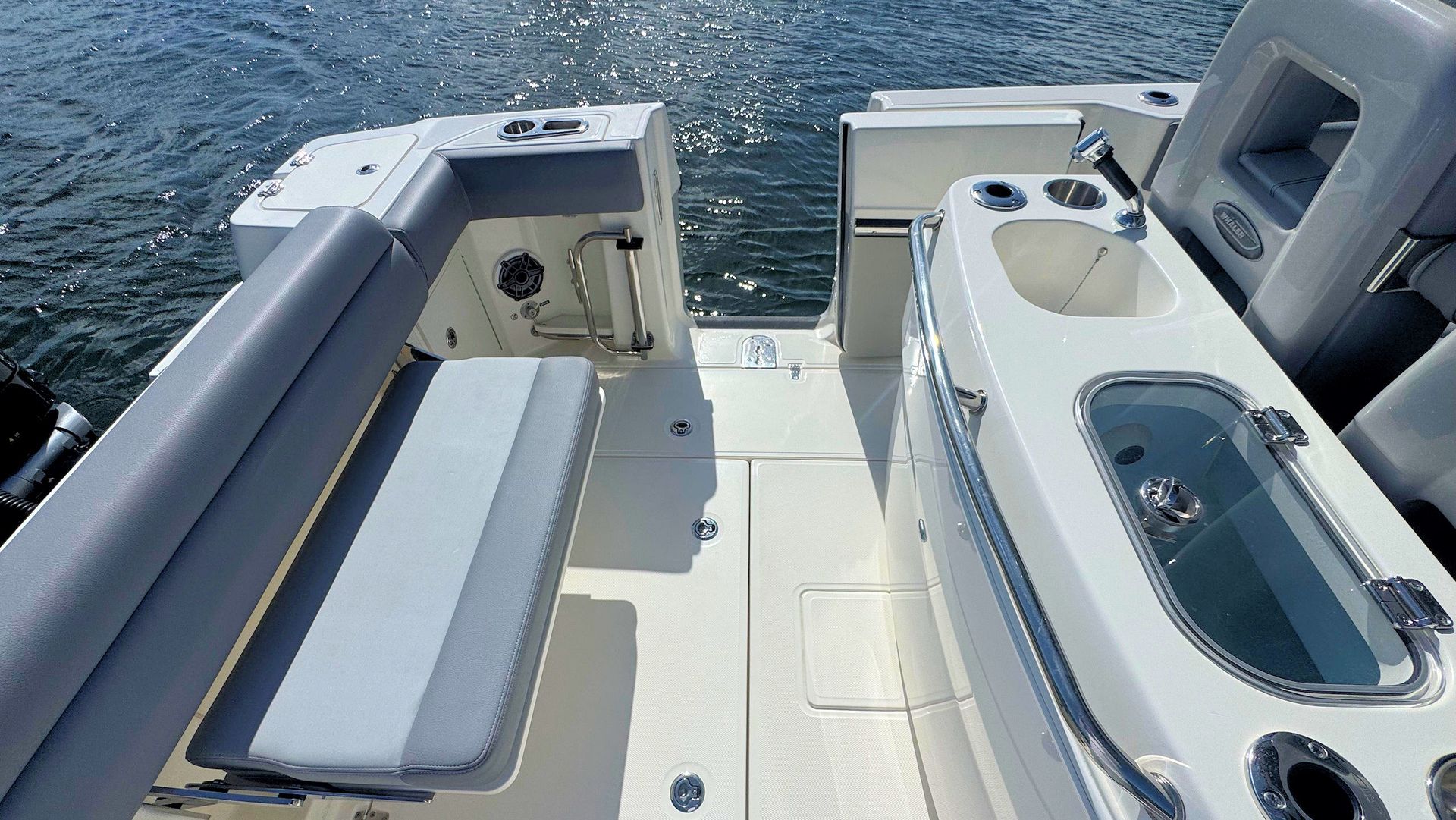 The rear deck of a boat with a gray upholstered bench seat, a white wet bar, a sink, and a livewell looking out to water.