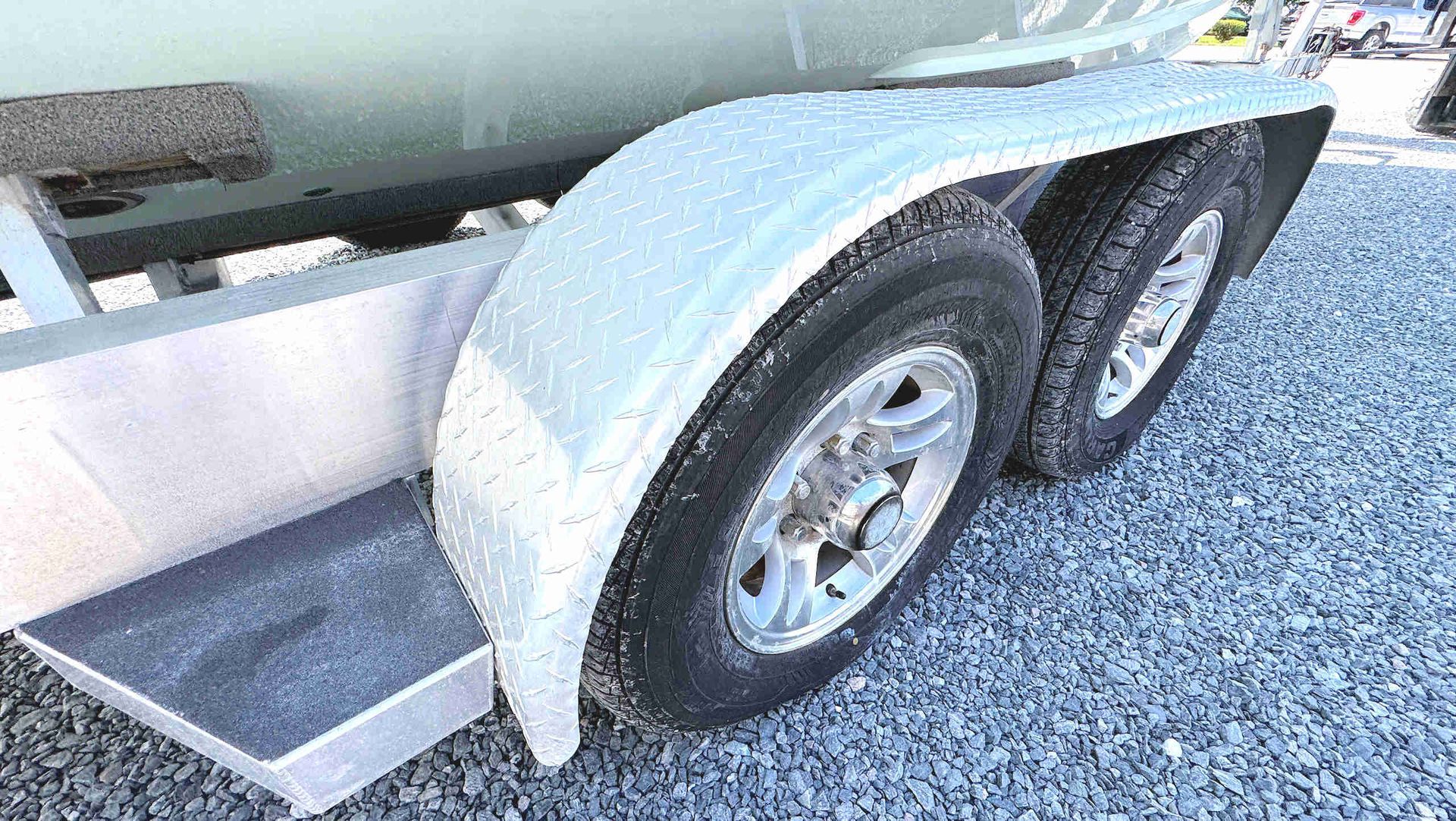 Aluminum boat trailer with tandem wheels and a small step, parked on gravel near a boat.