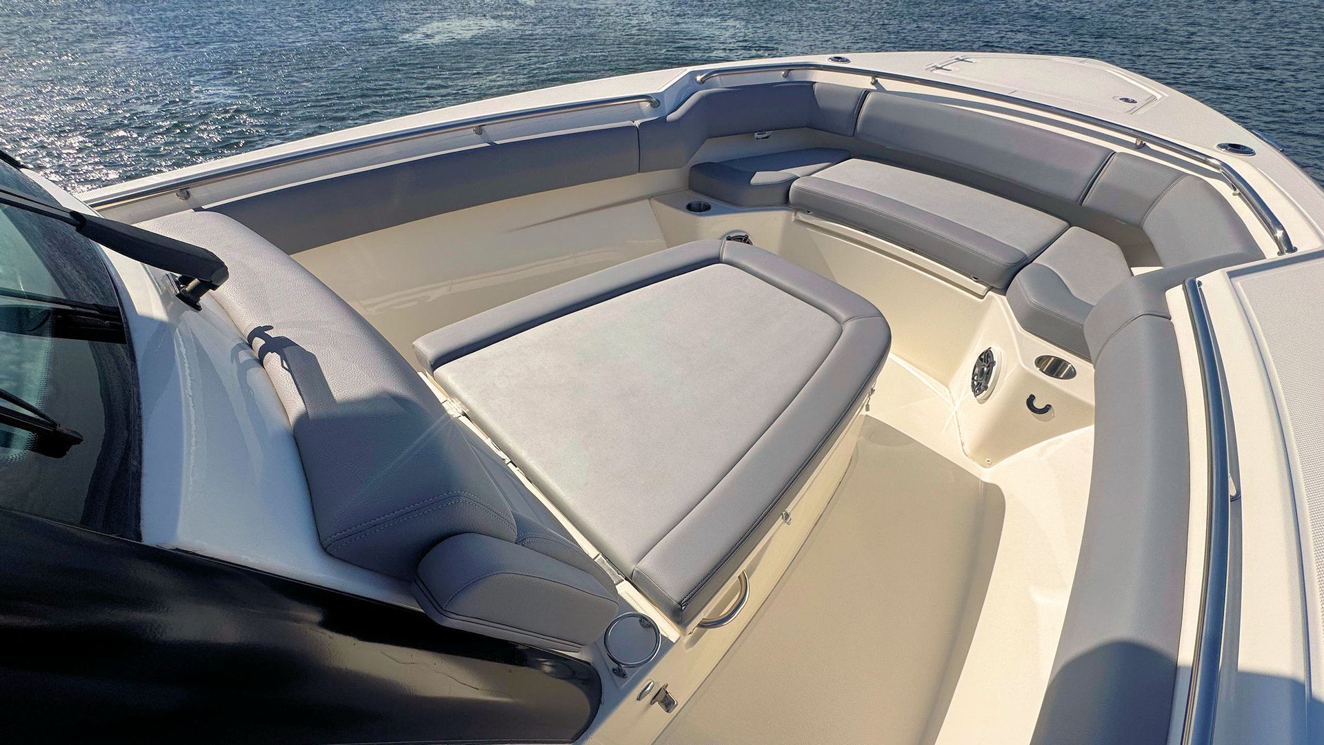 The bow area of a motorboat features cushioned gray seating, a central lounge pad, and white decking on sunny water.