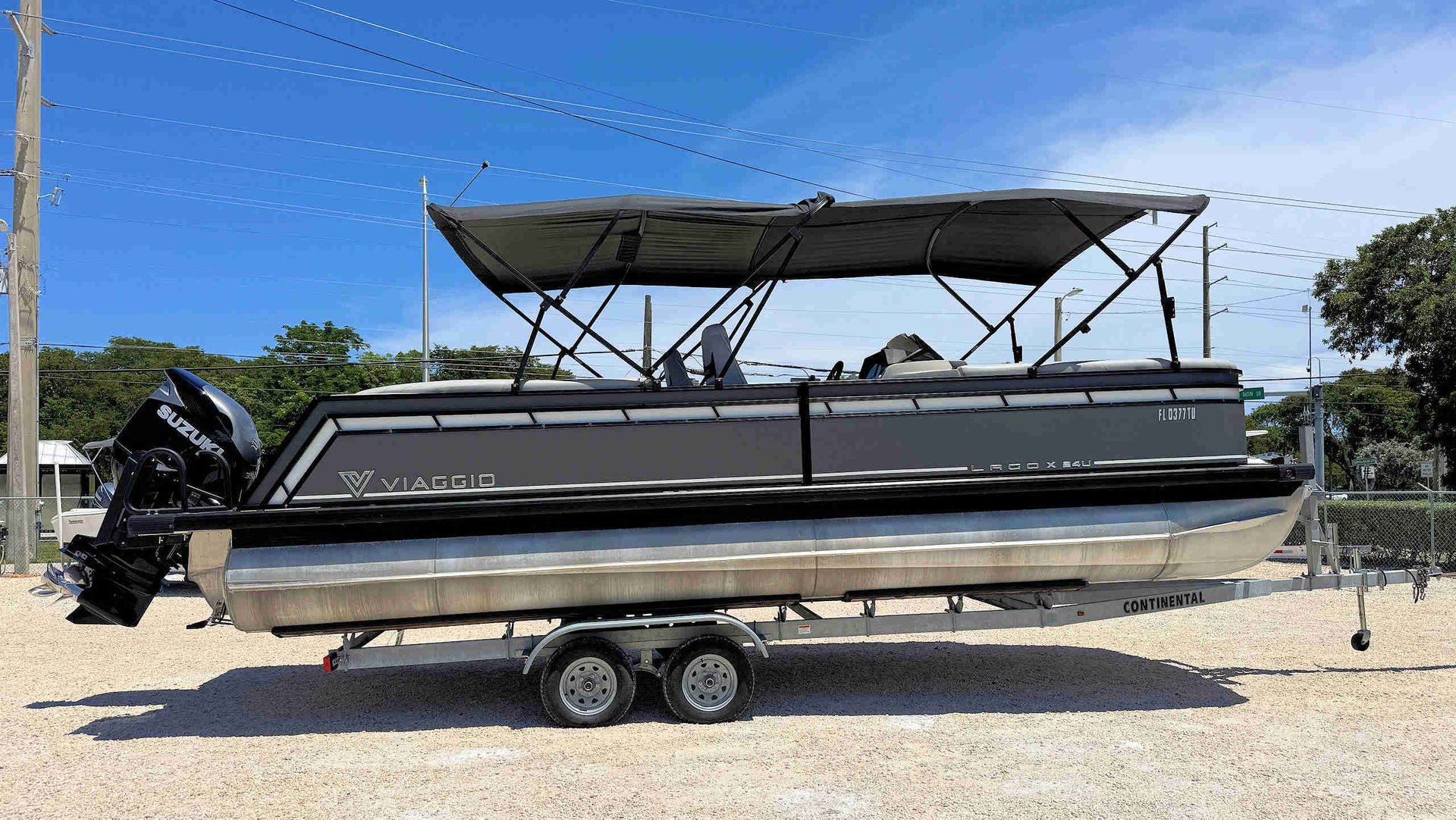 Black tritoon boat on a trailer with a canopy, parked on gravel under a blue sky.