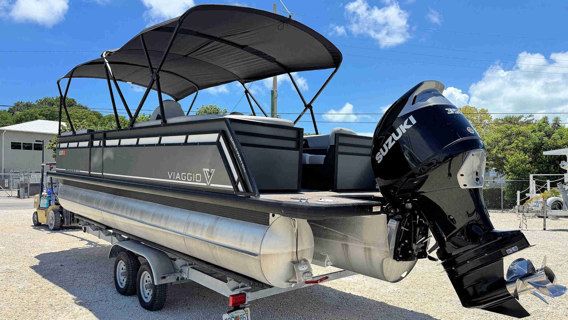 Tritoon boat on a trailer with black bimini tops and outboard motor, parked outdoors under a blue sky