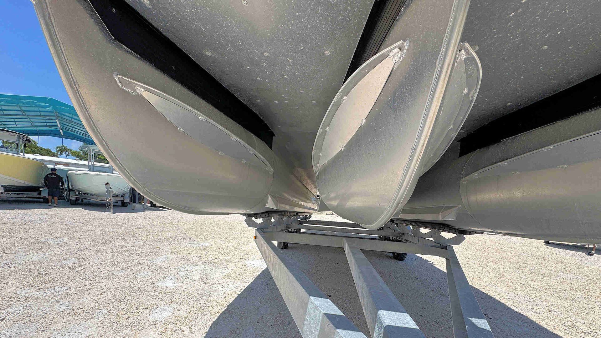 Tritoon boat hulls resting on a trailer over gravel, viewed from below outdoors on a sunny day