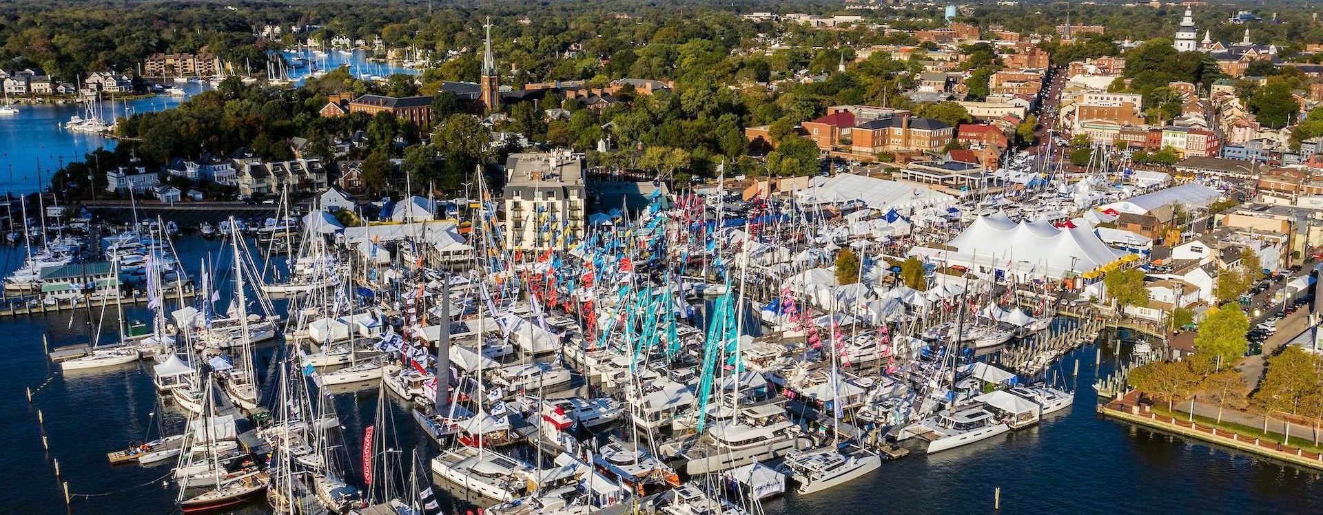 Annapolis Sailboat Show Aerial view of a boat show with numerous sailboats and tents on the water. Buildings and trees are in the background.
