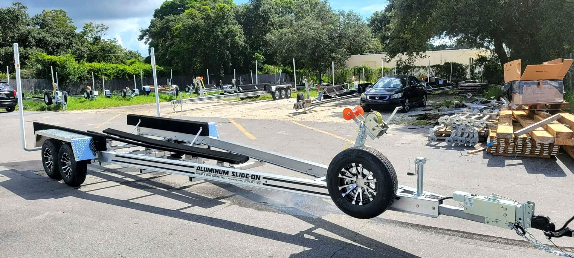 A boat trailer on asphalt. Trees, a building, and debris are in the background.