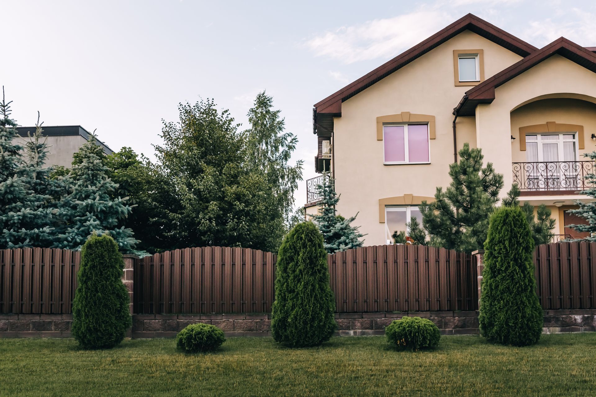 Tan house with brown fence, green trees, and bushes in front.