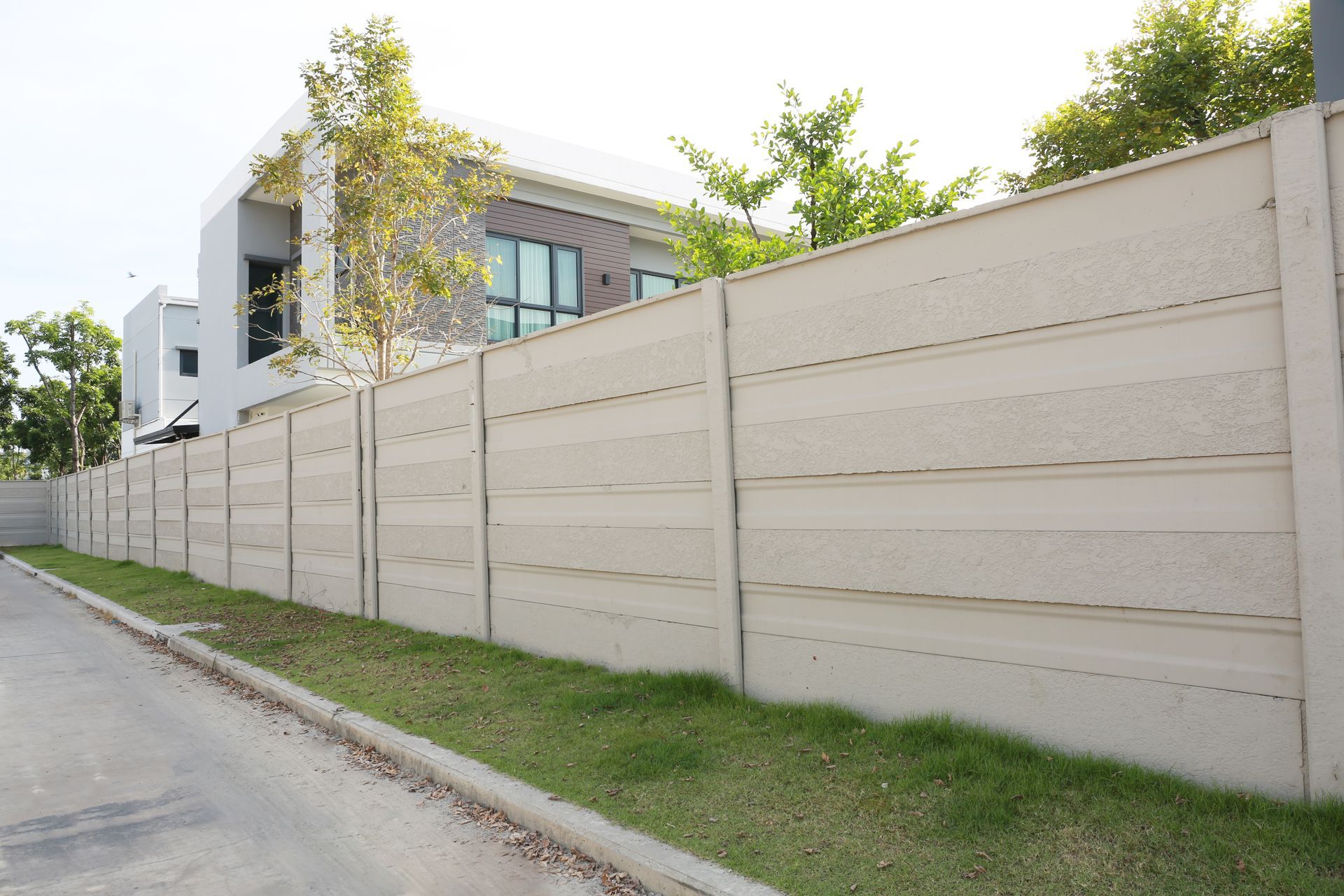 A gray concrete fence borders a residential property with a modern house, grass, and a street.