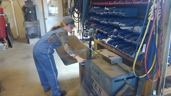 Woman in blue overalls lifting metal object in a workshop, shelves of blue bags in background.