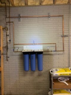 Water filtration system with three blue filters and copper pipes mounted on a brick wall.