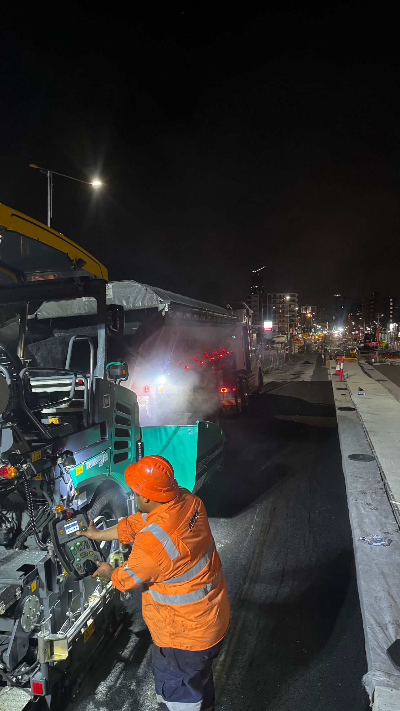 Worker in orange safety gear repairs machinery at night; steam visible.