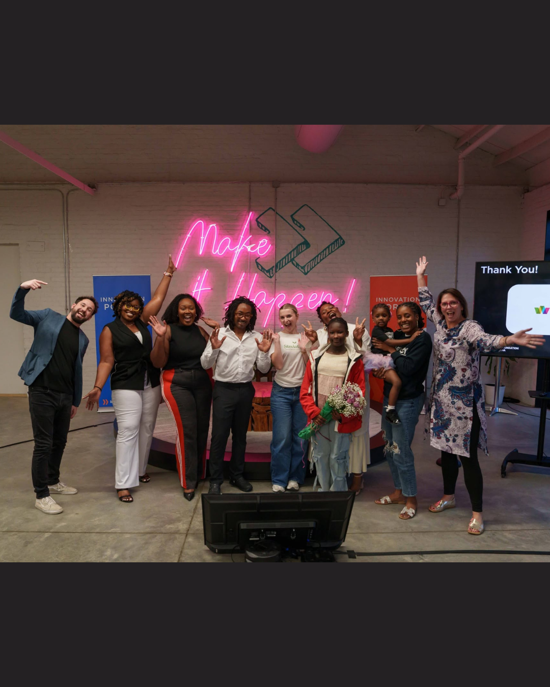 Group of people posing onstage under pink neon “Make it Happen” sign, smiling in a lively event setting