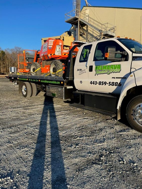 White flatbed truck hauling an orange utility lift on a gravel lot under a clear blue sky
