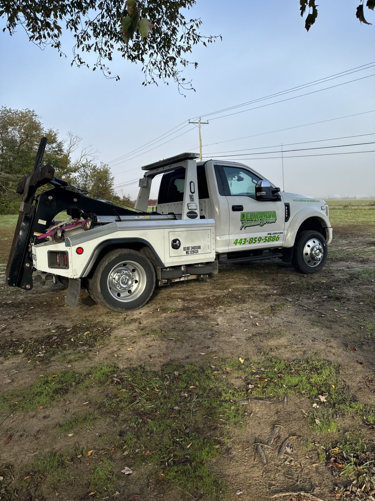 White tow truck parked on a dirt slope near trees and power lines.