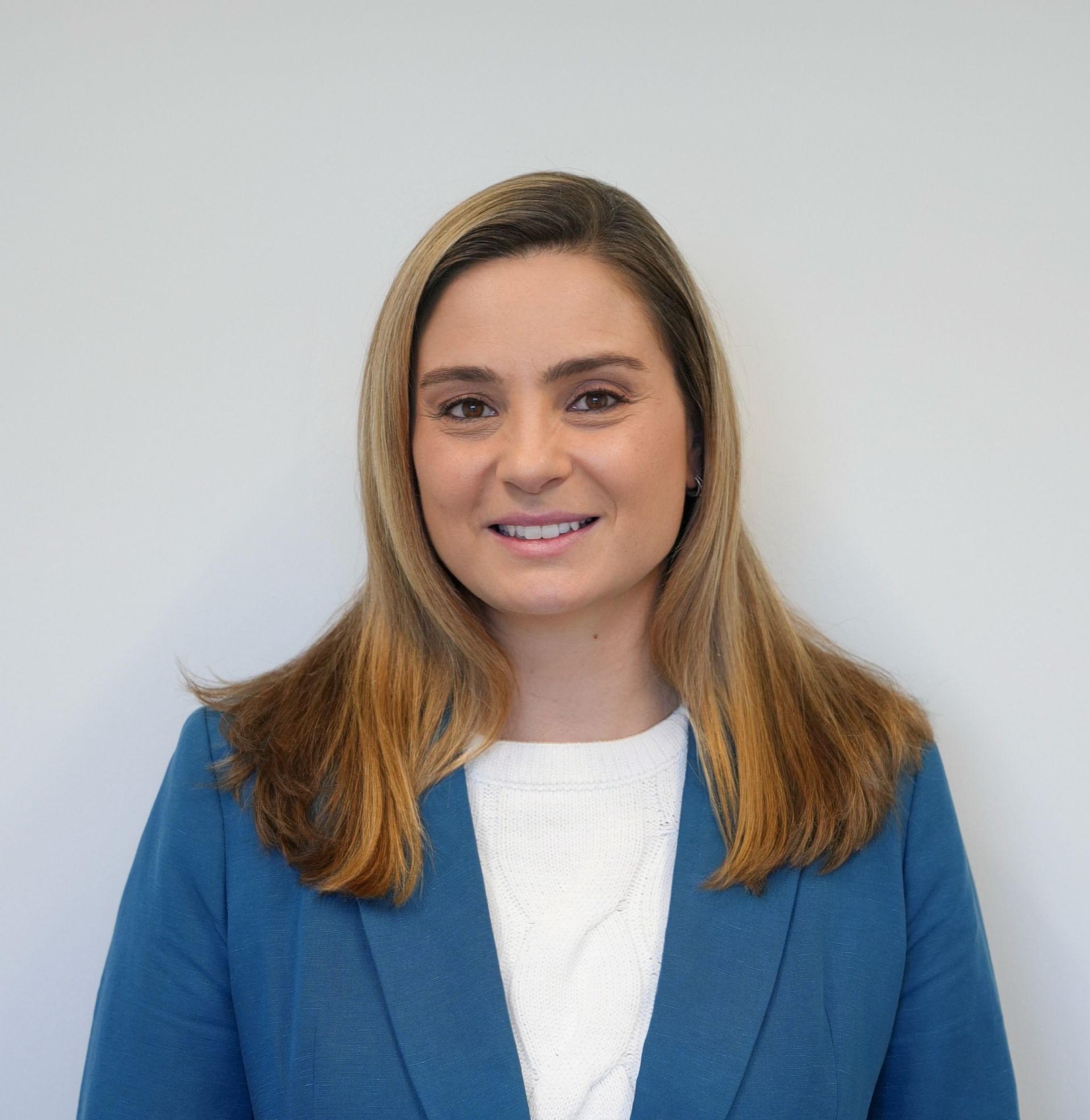 Woman with long, light brown hair wearing a blue blazer over a white sweater, smiling against a white backdrop.