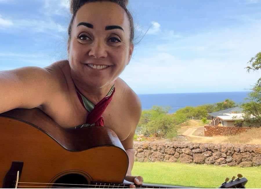 A woman is smiling while holding a guitar in front of the ocean.