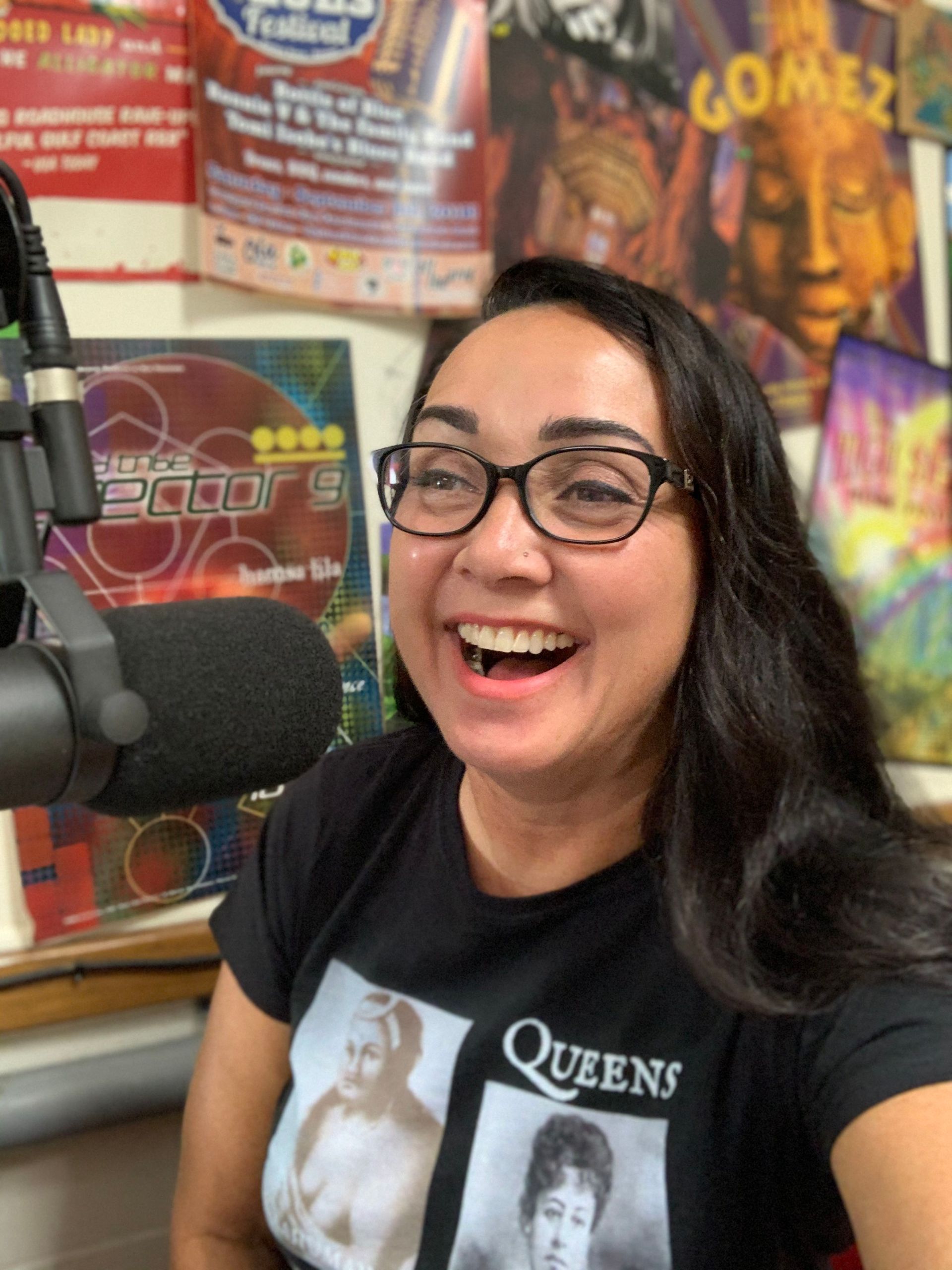 A woman wearing glasses and a queens shirt smiles in front of a microphone