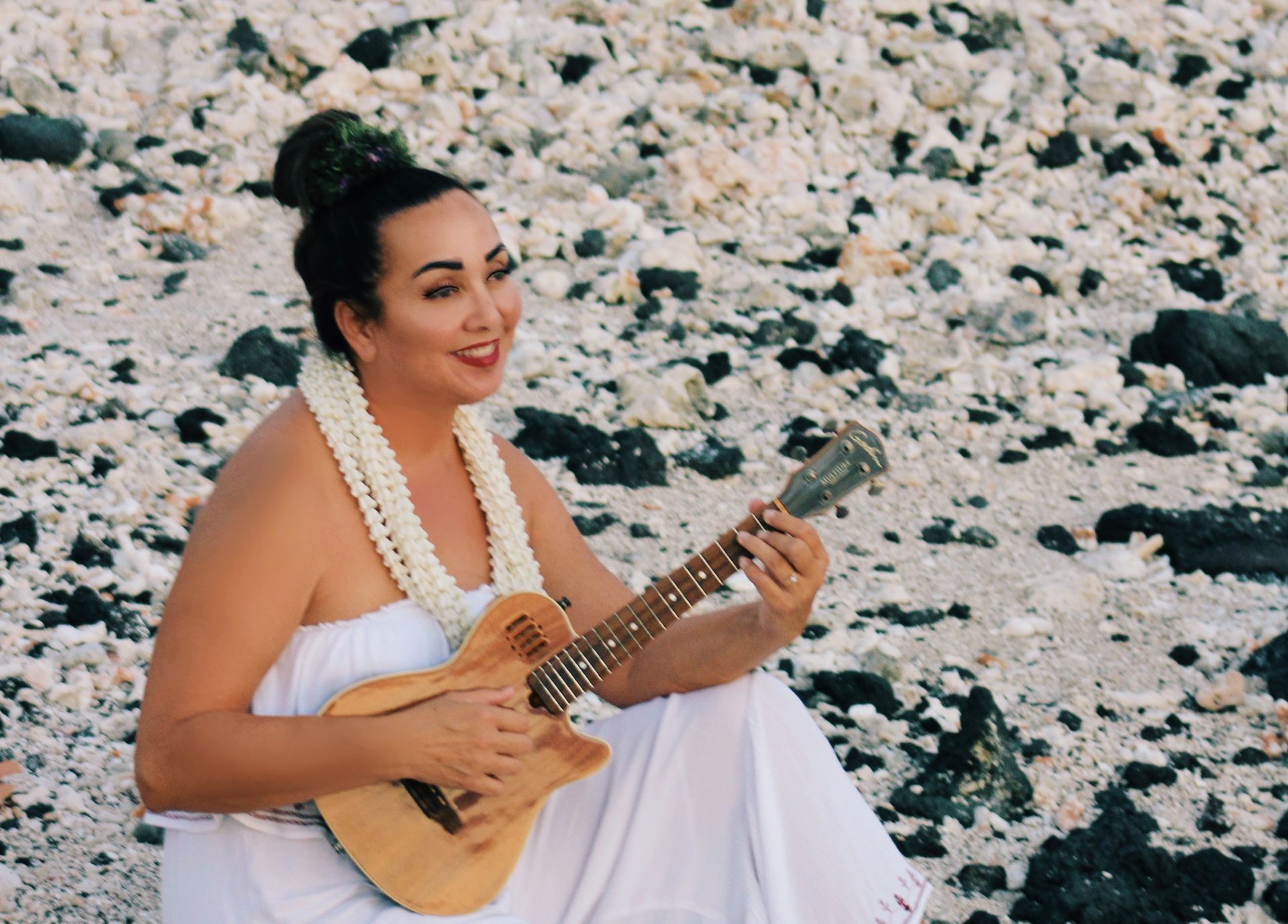 A woman in a white dress is sitting on the beach playing a guitar.