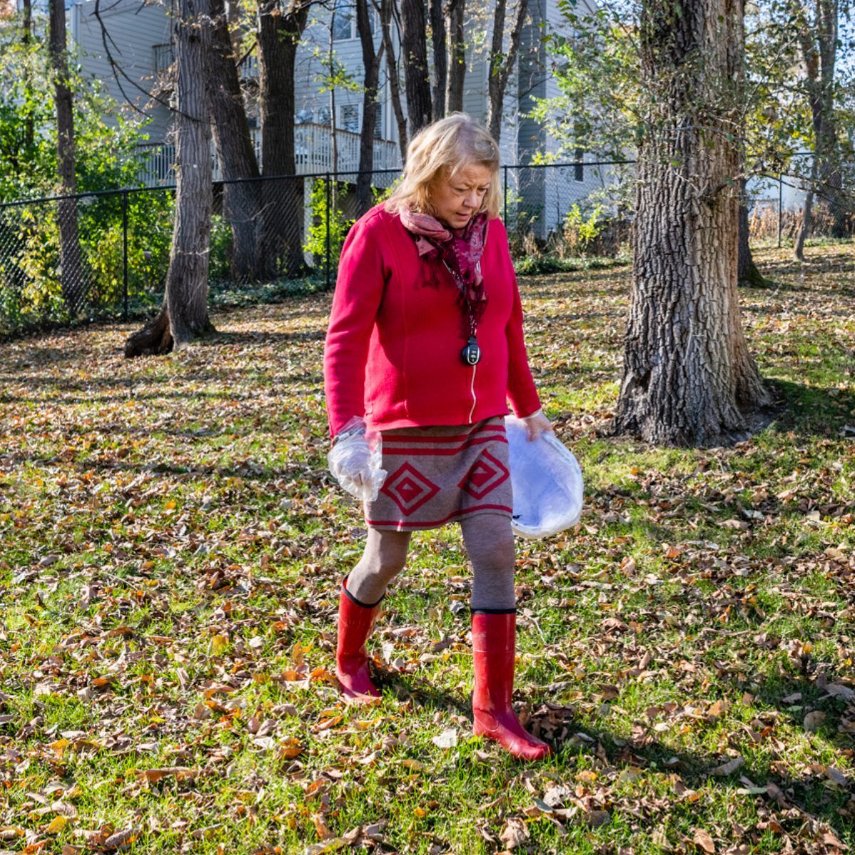 Woman in red jacket and boots, walking outdoors holding a white object in a wooded area with leaves.