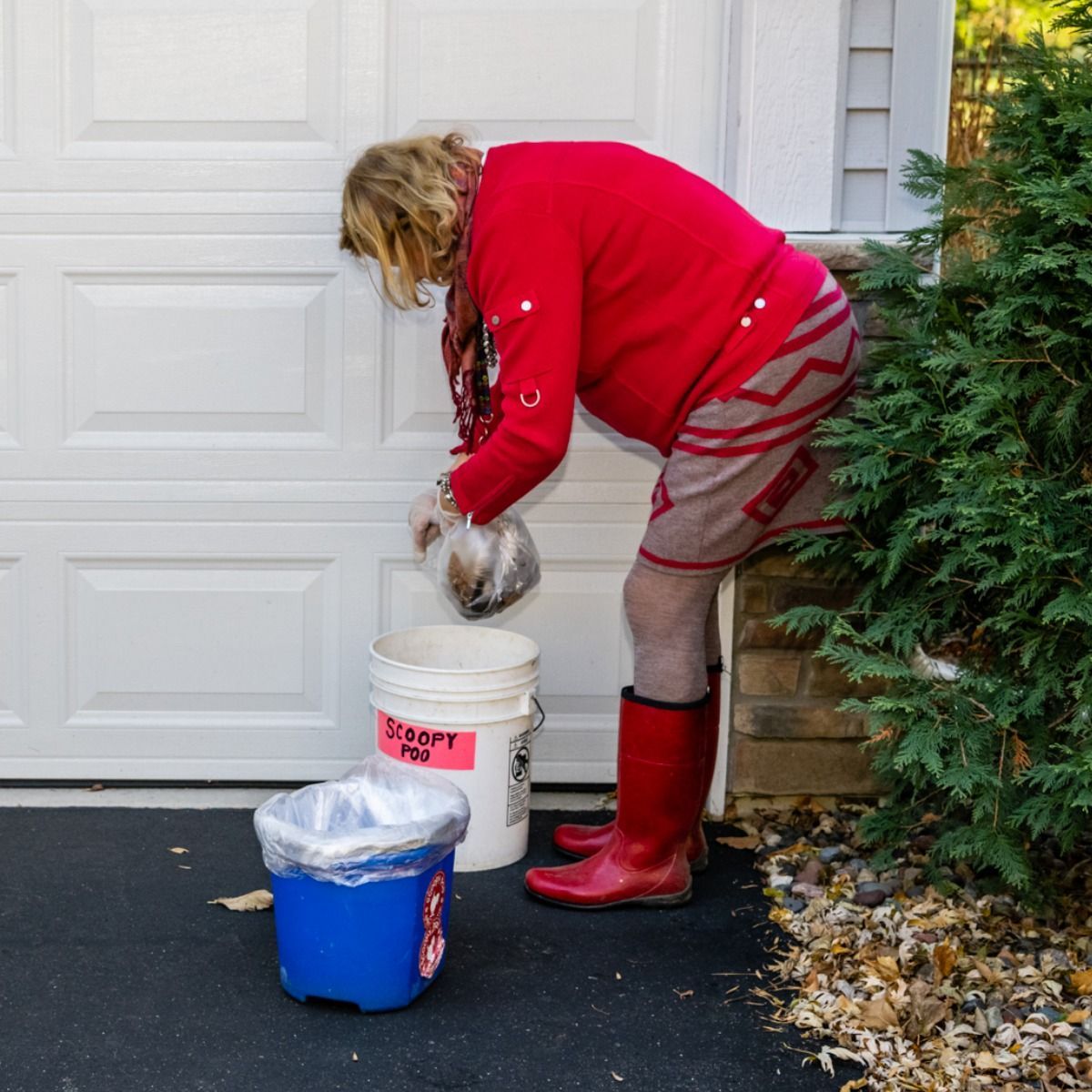 Person in red sweater and boots putting something into a white bucket next to a garage door.
