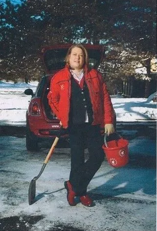 Person in red jacket and shoes stands by red car, holding bucket and leaning on shovel, in a snowy setting.