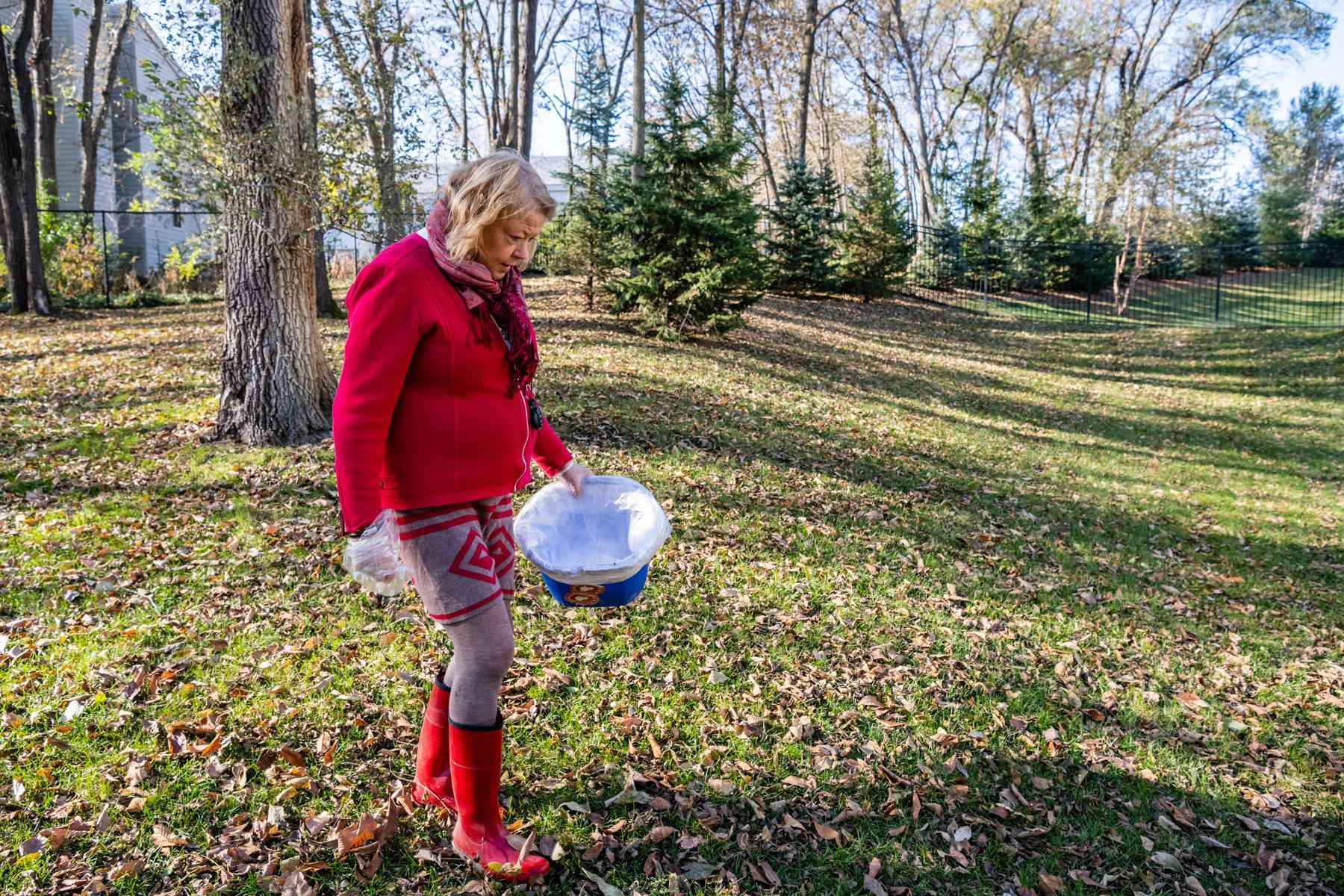 Woman in red jacket and boots holding a white and blue bowl in a grassy outdoor area, looking down.