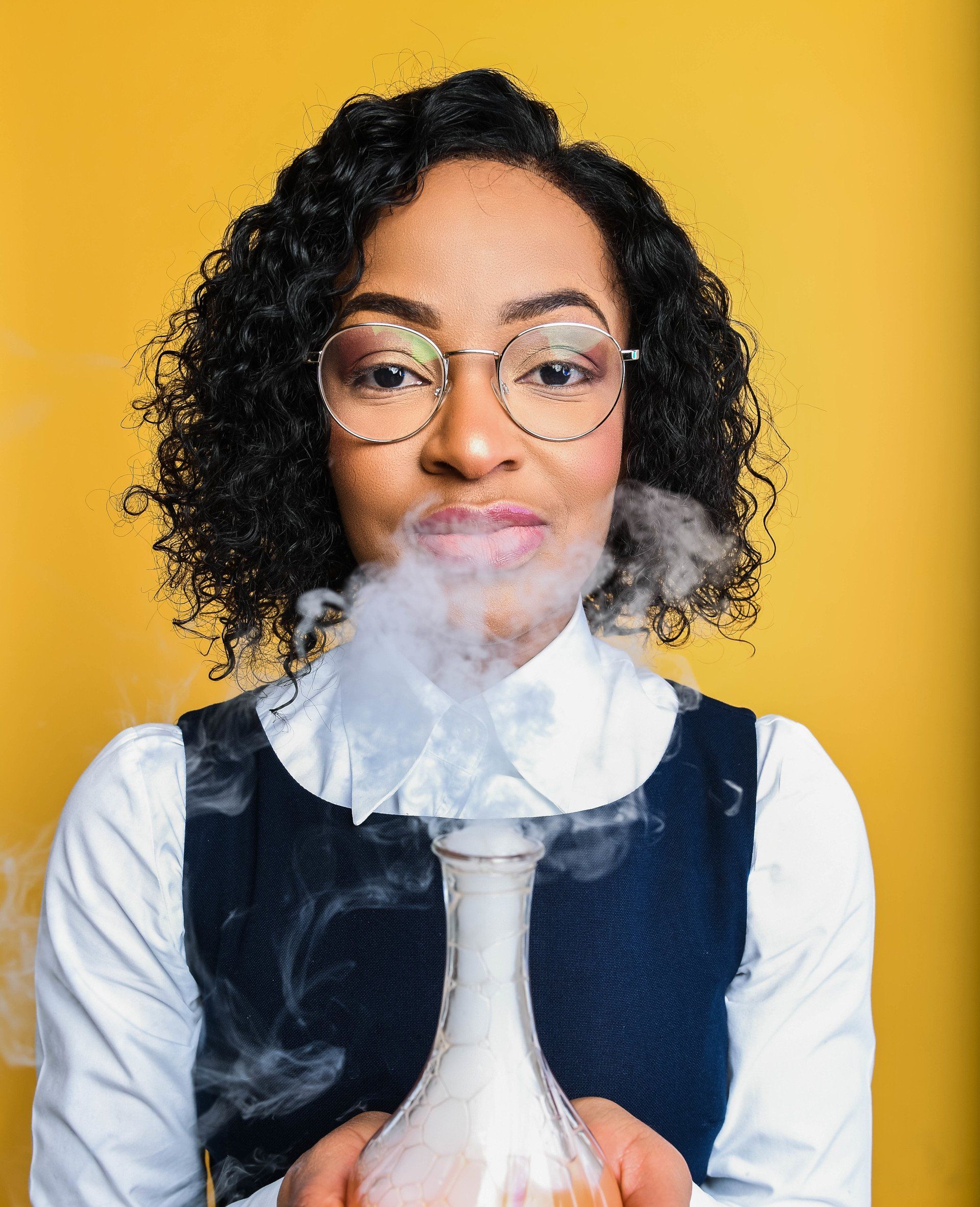 A woman wearing glasses is holding a beaker with smoke coming out of it.