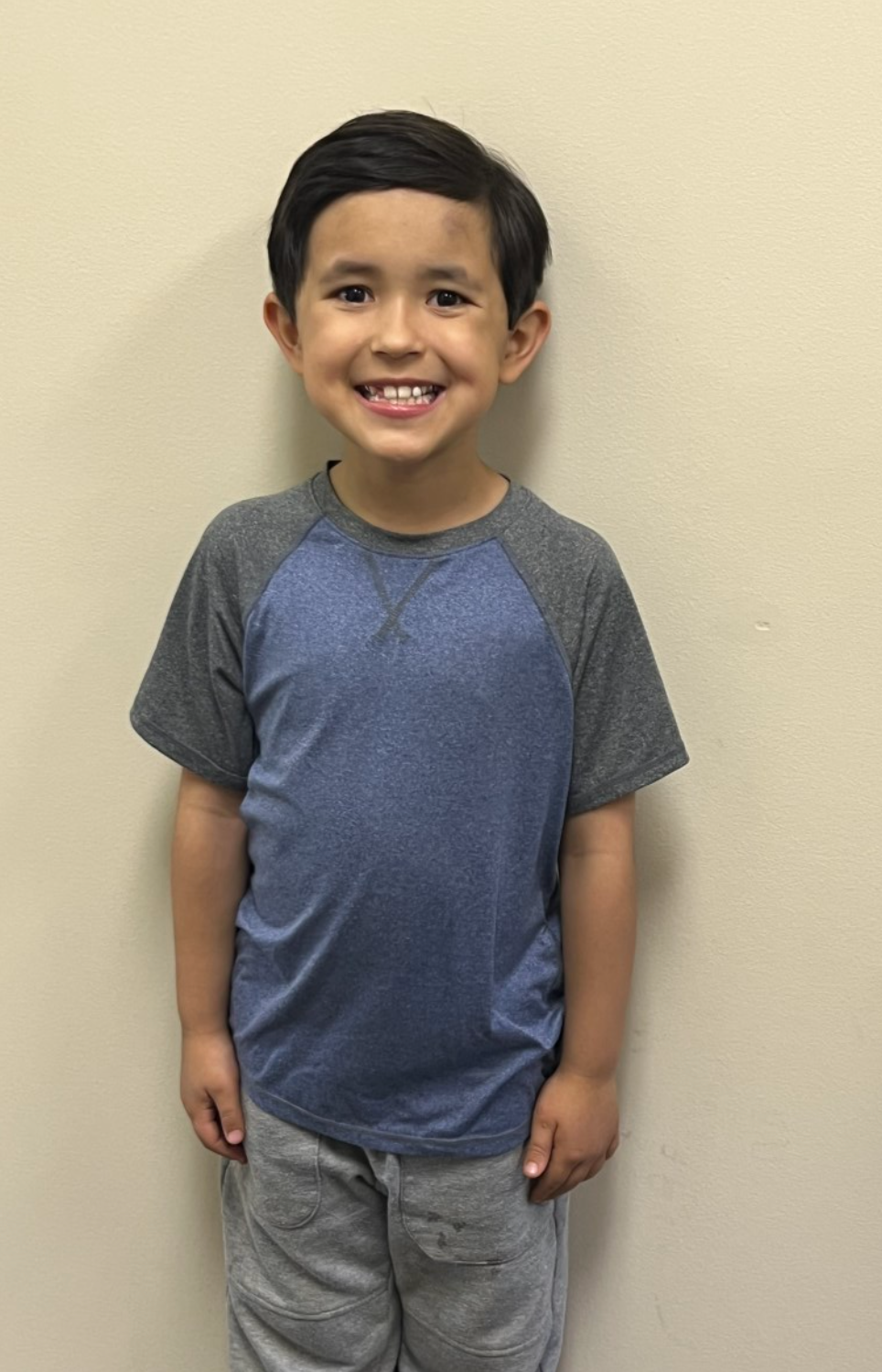 A young boy (student) wearing a blue shirt and grey sweatpants is smiling while standing in front of a wall.