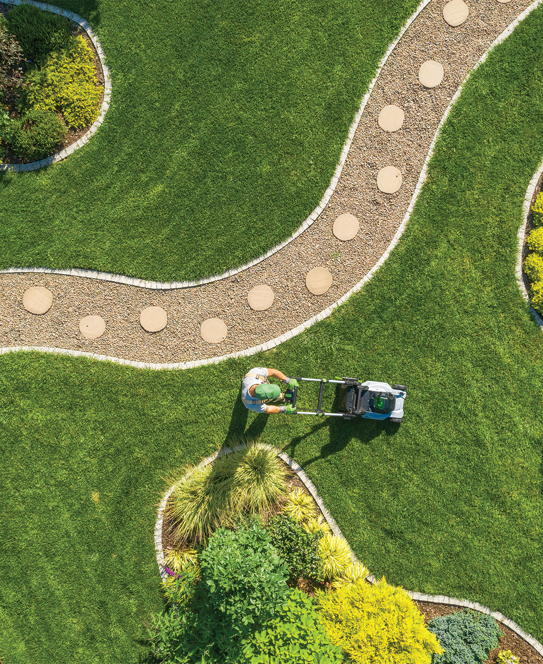 Person mowing green lawn with a pathway of stepping stones and garden beds.