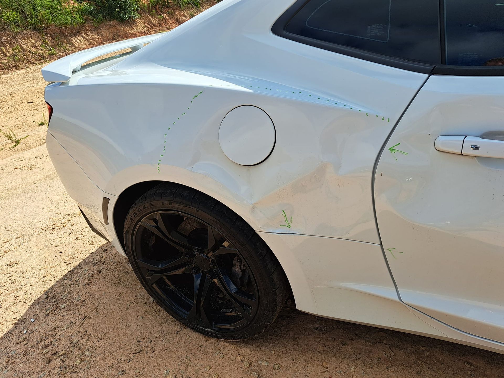A white car with a damaged fender is parked on a dirt road.