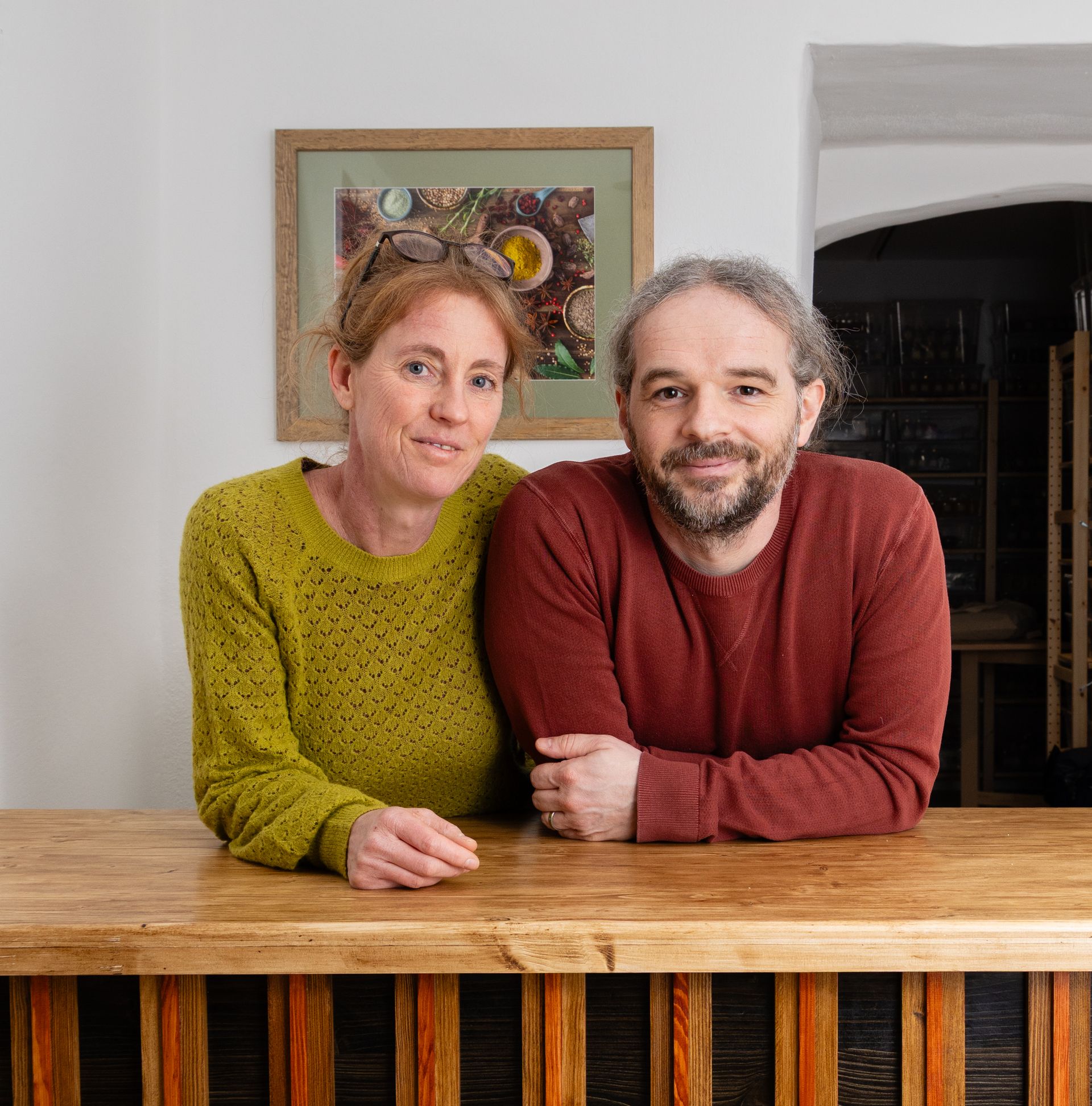 Eine Frau in einem grünen Pullover und ein Mann in einem roten Pullover stehen nebeneinander und lehnen sich in einem Innenraum an eine hölzerne Theke.