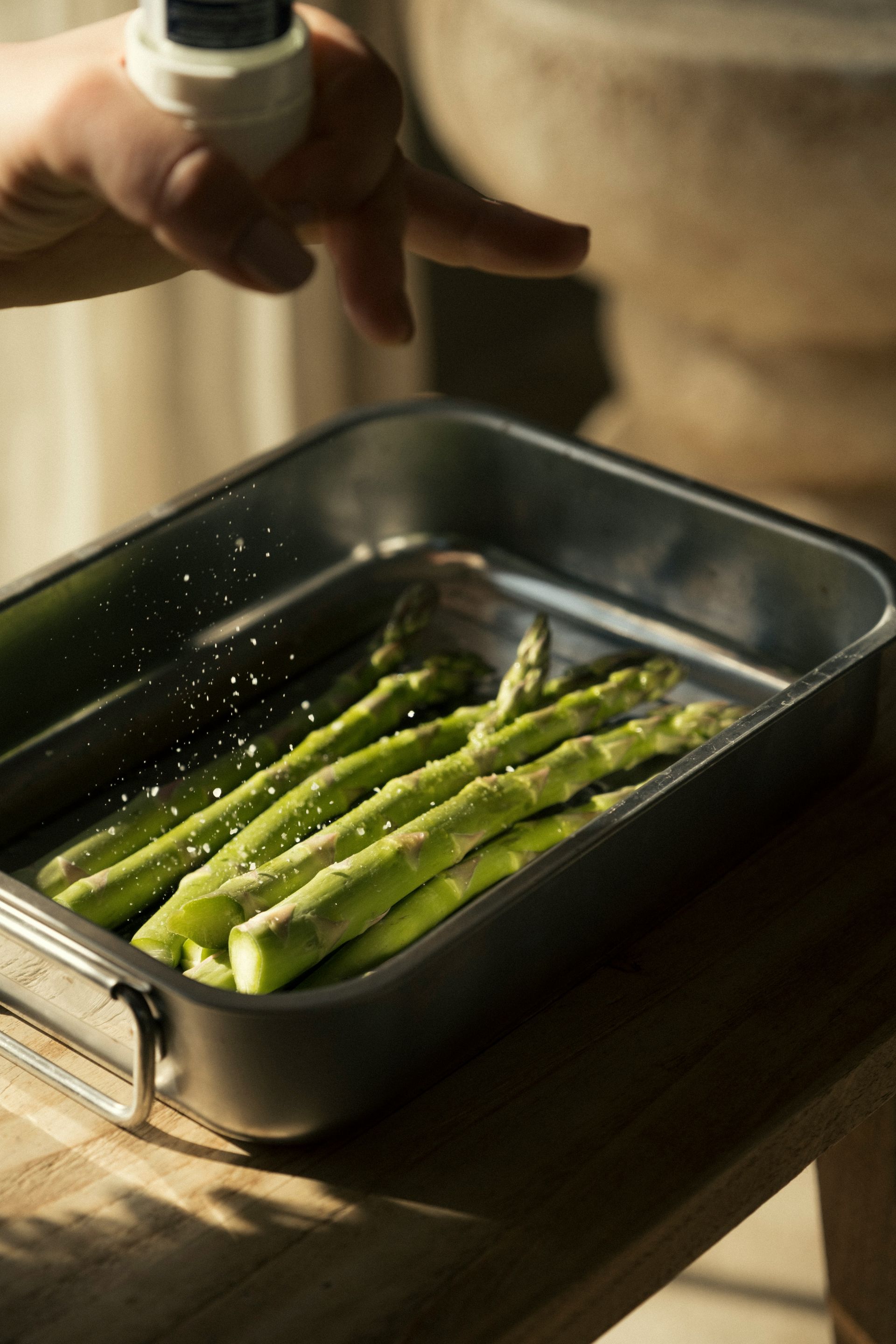 Eine Person bestreut mit der Hand frische grüne Spargelspitzen in einer Metallbackform auf einem Holztisch mit Gewürzen.
