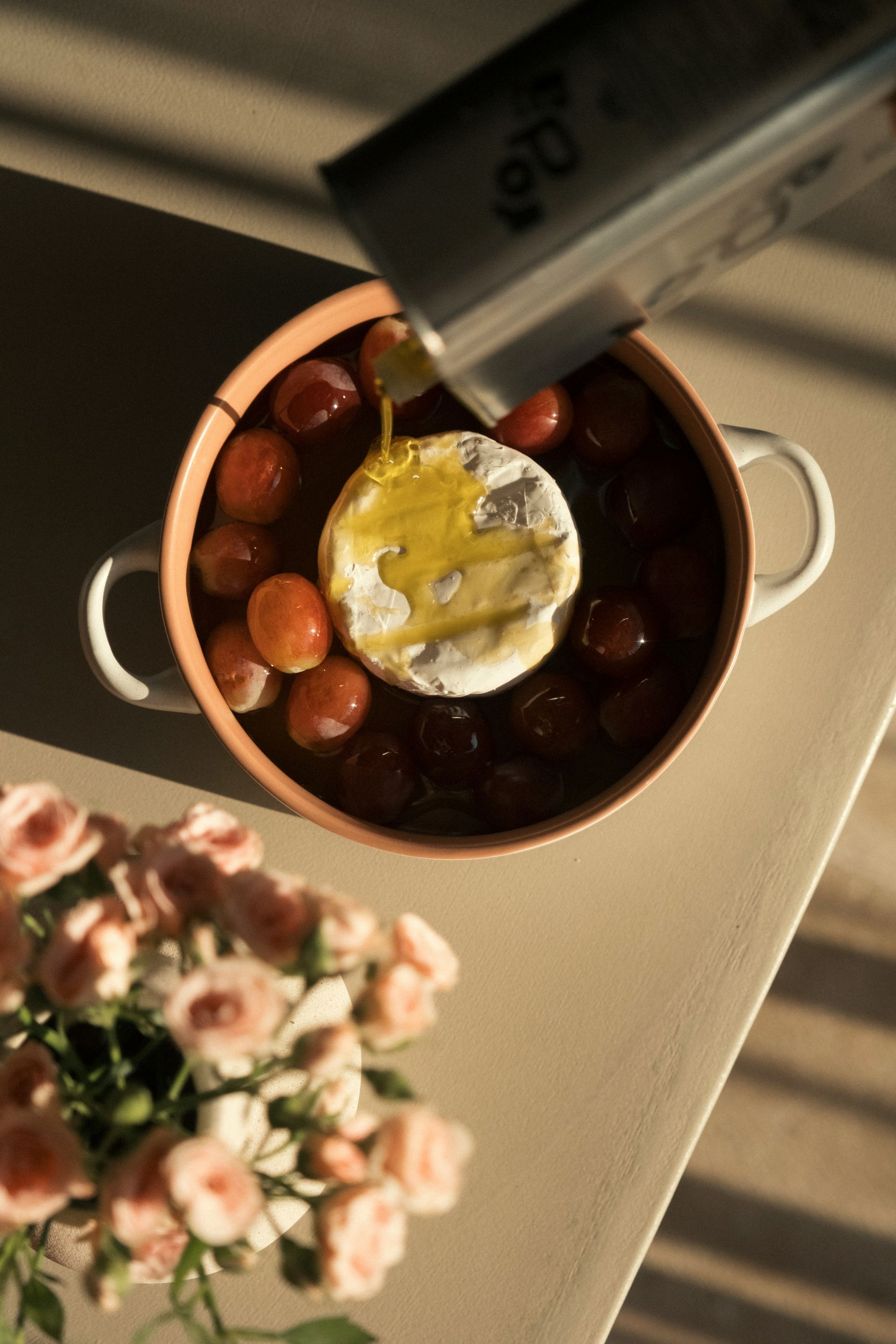 In einer Keramikschale wird Olivenöl über einen runden Brie-Käse gegossen, der von Weintrauben umgeben ist; im Vordergrund sind Rosen zu sehen.