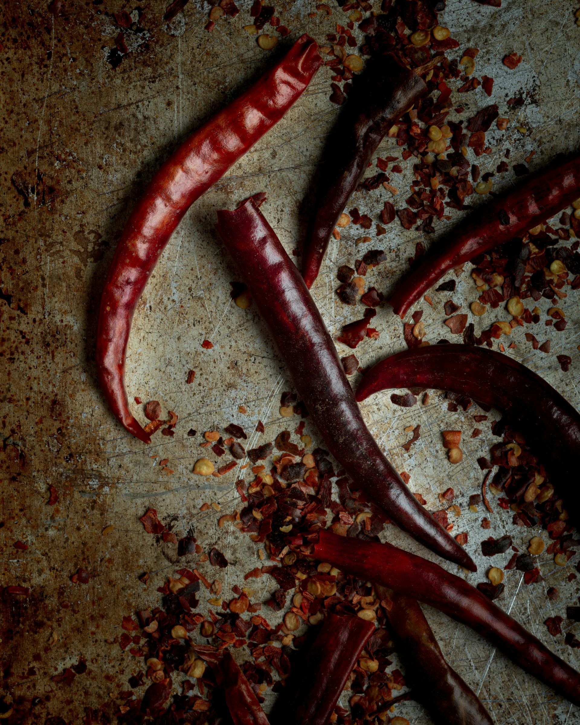 Getrocknete rote Chilischoten und verstreute Chiliflocken auf einem strukturierten, dunkelmetallischen Hintergrund.