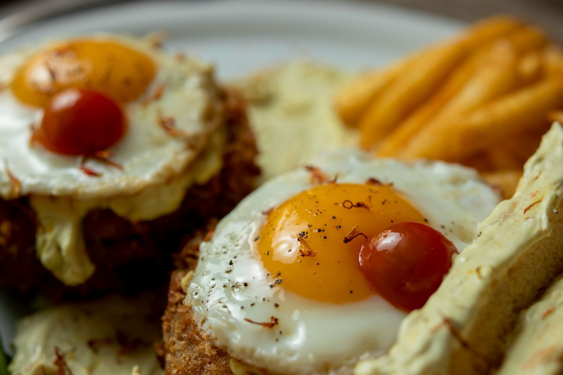 Zwei Spiegeleier, jeweils mit einer Kirschtomate belegt, serviert auf knusprigen Frikadellen mit einer dicken, cremigen Soße und Pommes frites.