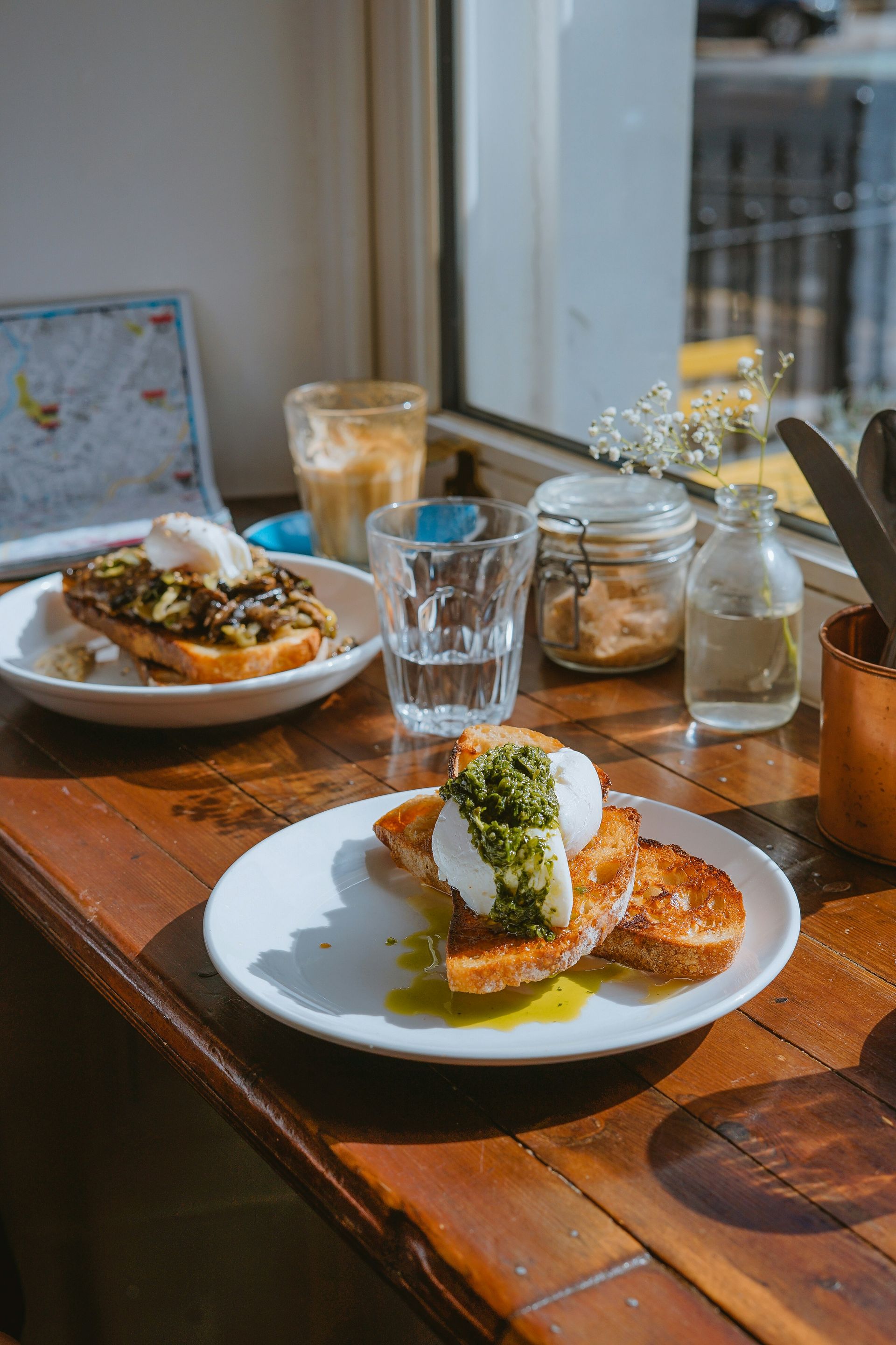 Zwei Teller mit pochierten Eiern auf Toast mit Kräutern, Kaffee und Wasser auf einem Holztisch an einem hellen Fenster.