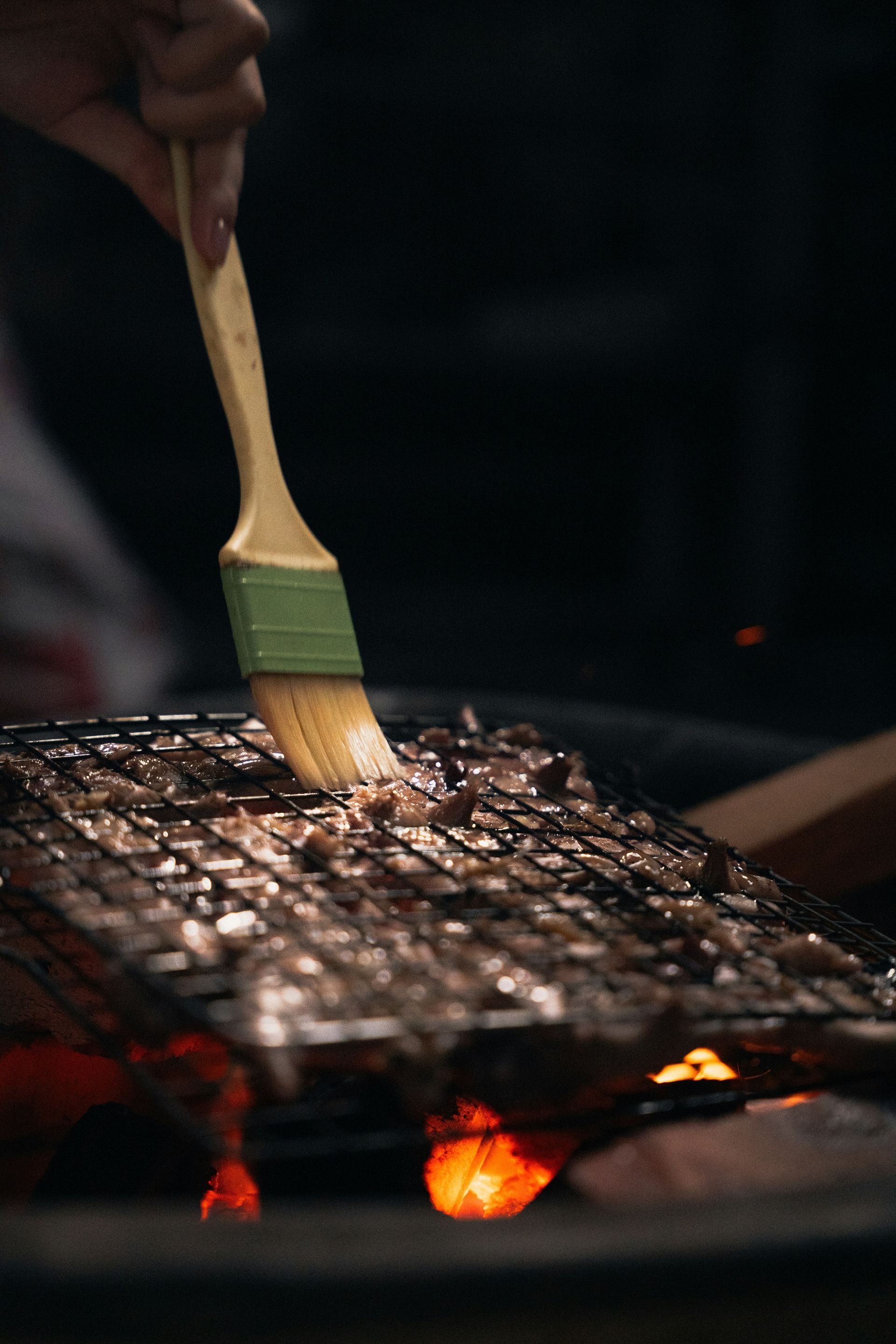 Eine Hand bestreicht mit einem grünen Silikonpinsel Fleisch, das auf einem Drahtgrill über glühender Holzkohle gart.