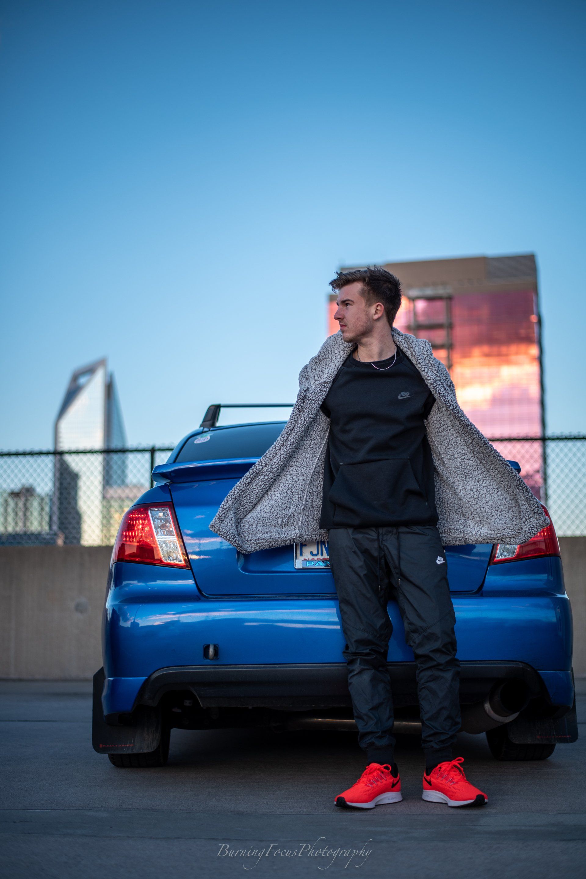 A man is standing in front of a blue car in a parking lot.