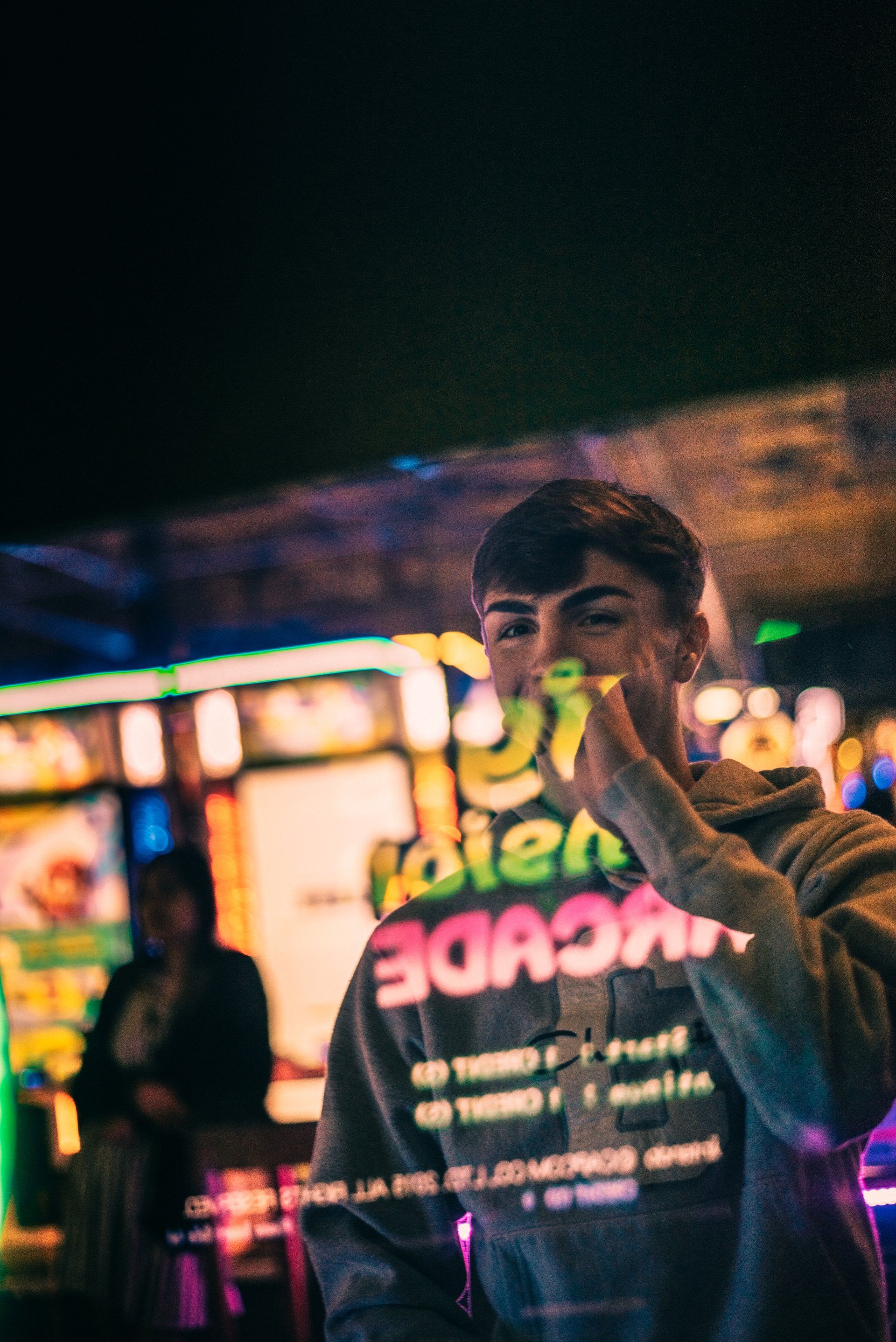 A man in a hoodie is standing in front of an arcade machine at night.