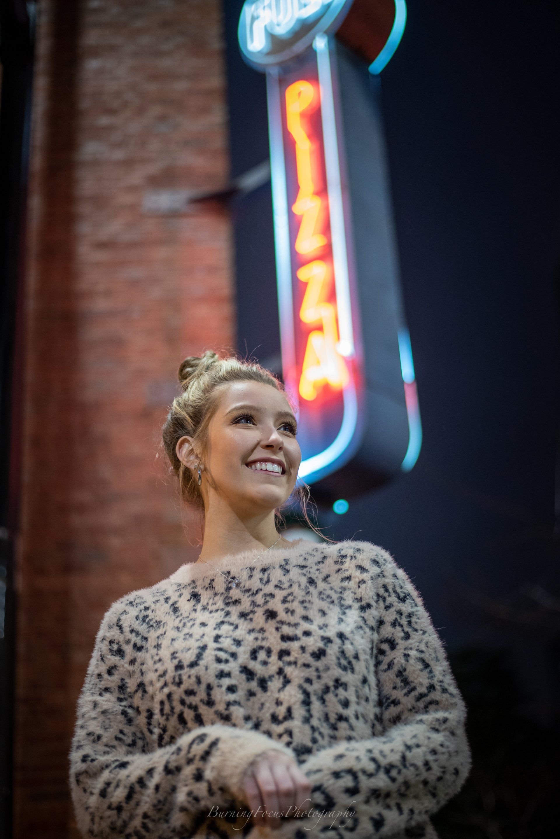 A woman in a leopard print sweater is standing in front of a neon pizza sign.