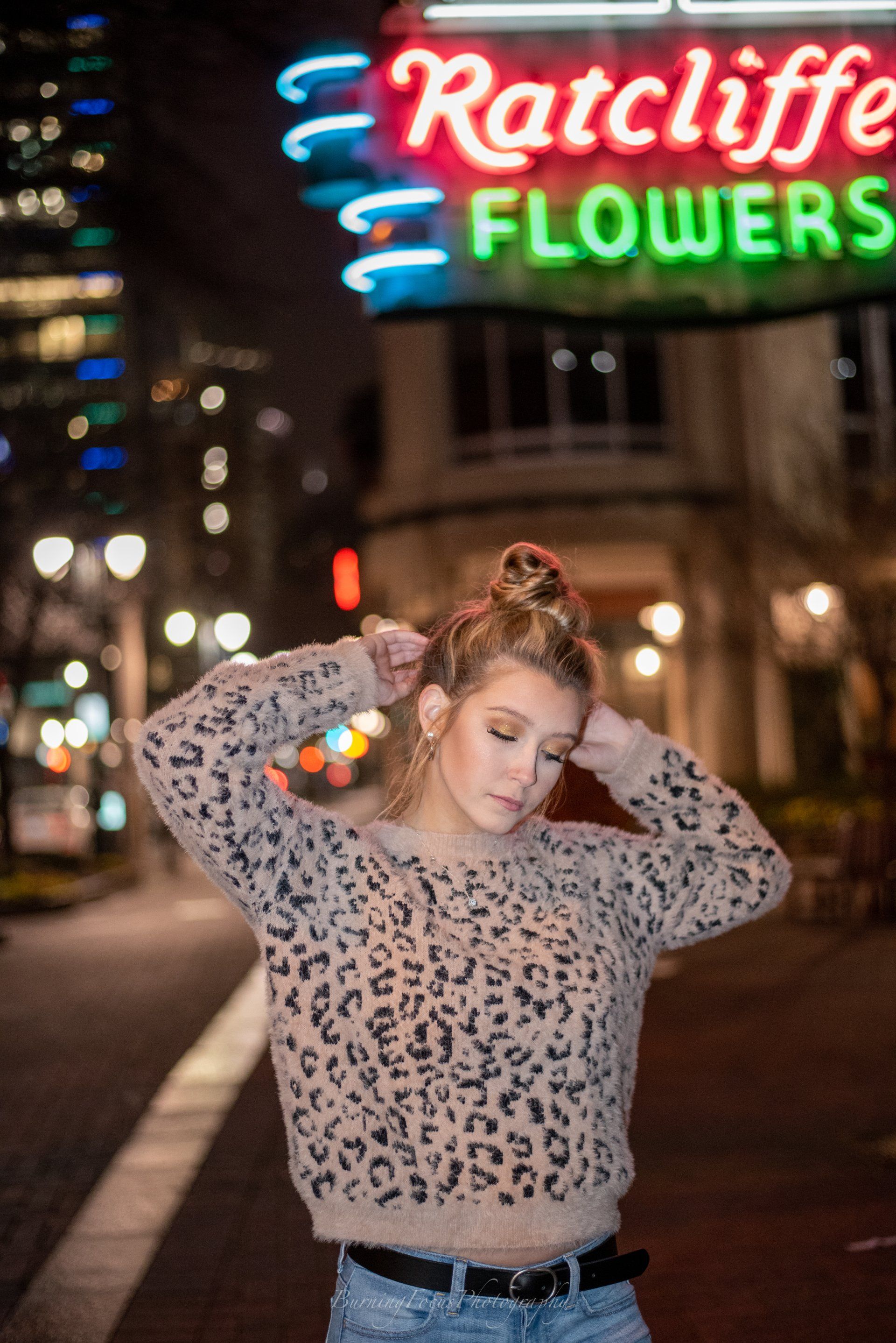 A woman in a leopard print sweater is standing in front of a neon sign for ratcliffe flowers.